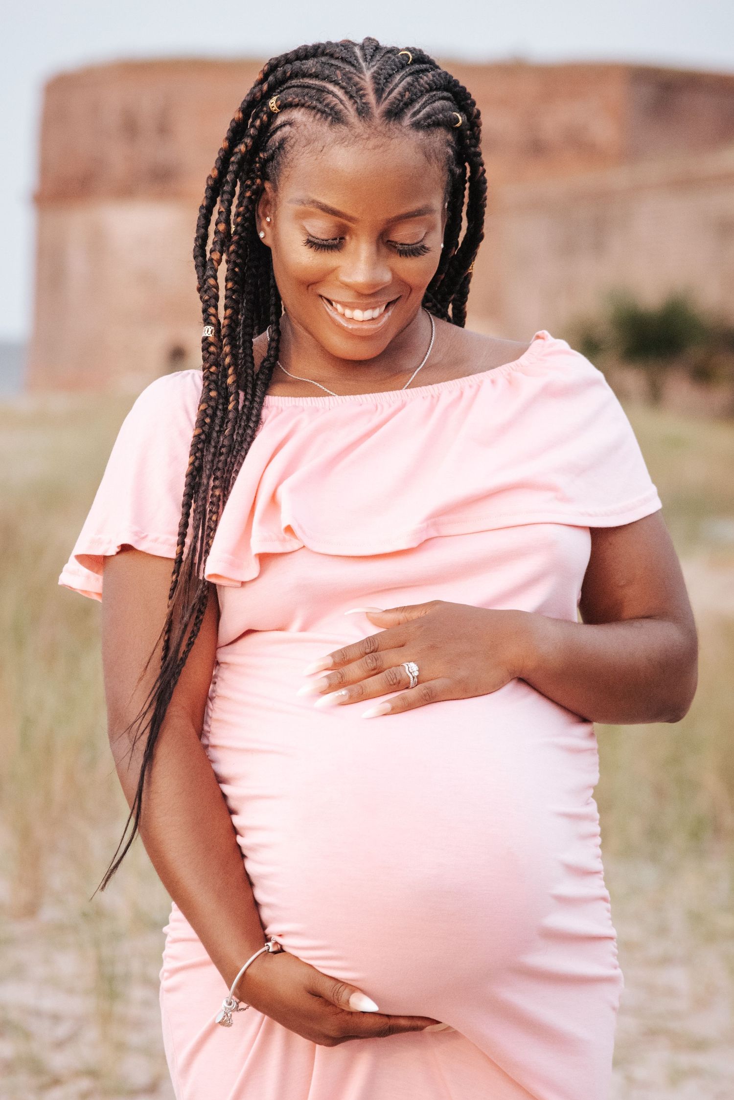 Glowing expectant mother in a pink off-shoulder dress stands outdoors with braided hair and gentle smile.