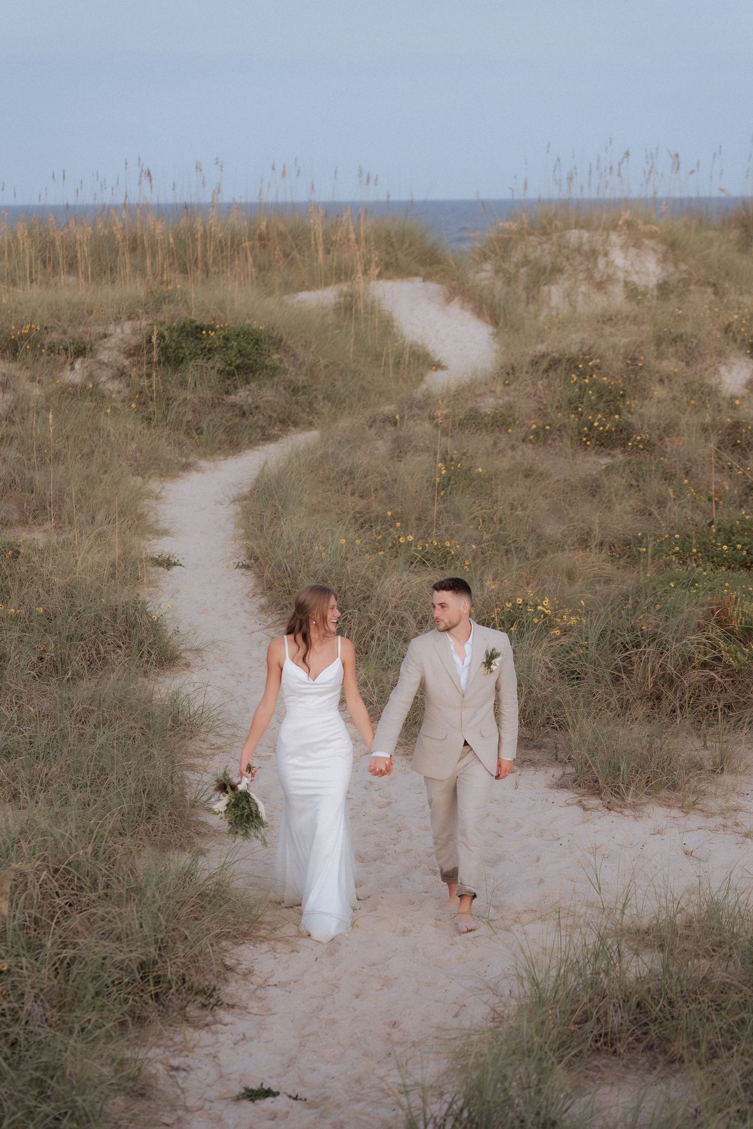 A couple walks hand in hand along a winding sandy beach path through coastal dunes and sea grass at sunset.