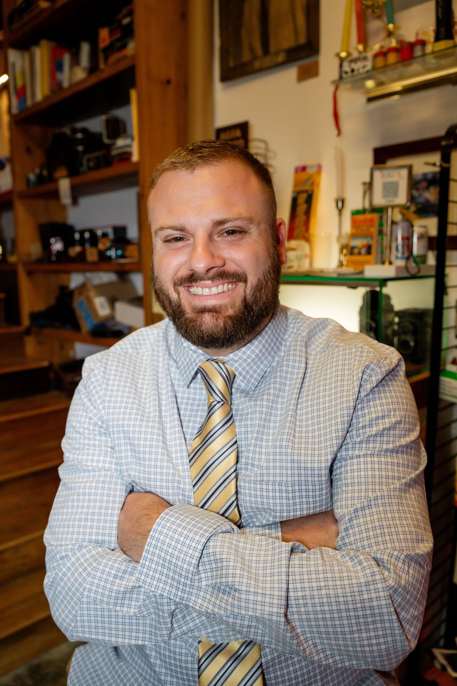 Smiling bearded man in checkered blazer and striped tie poses with arms crossed in a cozy shop filled with shelves.