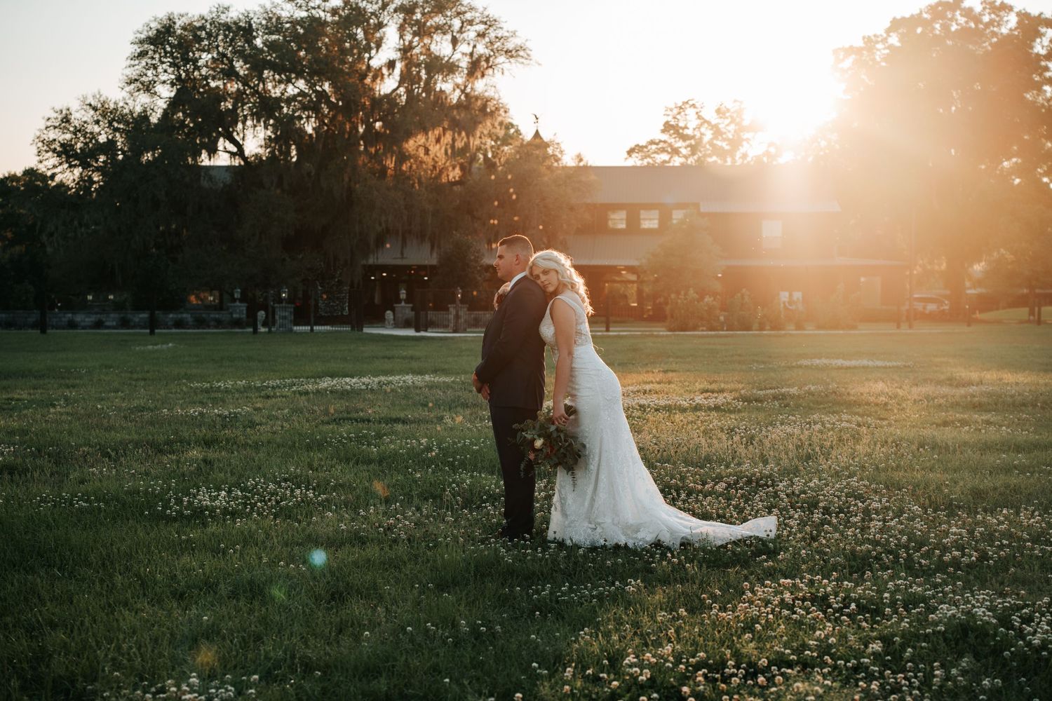 Two people in wedding attire stand together in a sunlit field at golden hour with a rustic building in the background.