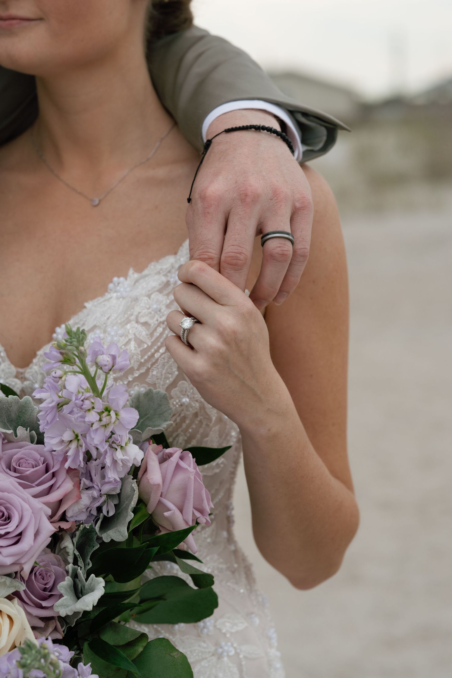 Bride and groom showing wedding rings, bride holding purple rose bouquet in lace dress outdoors.