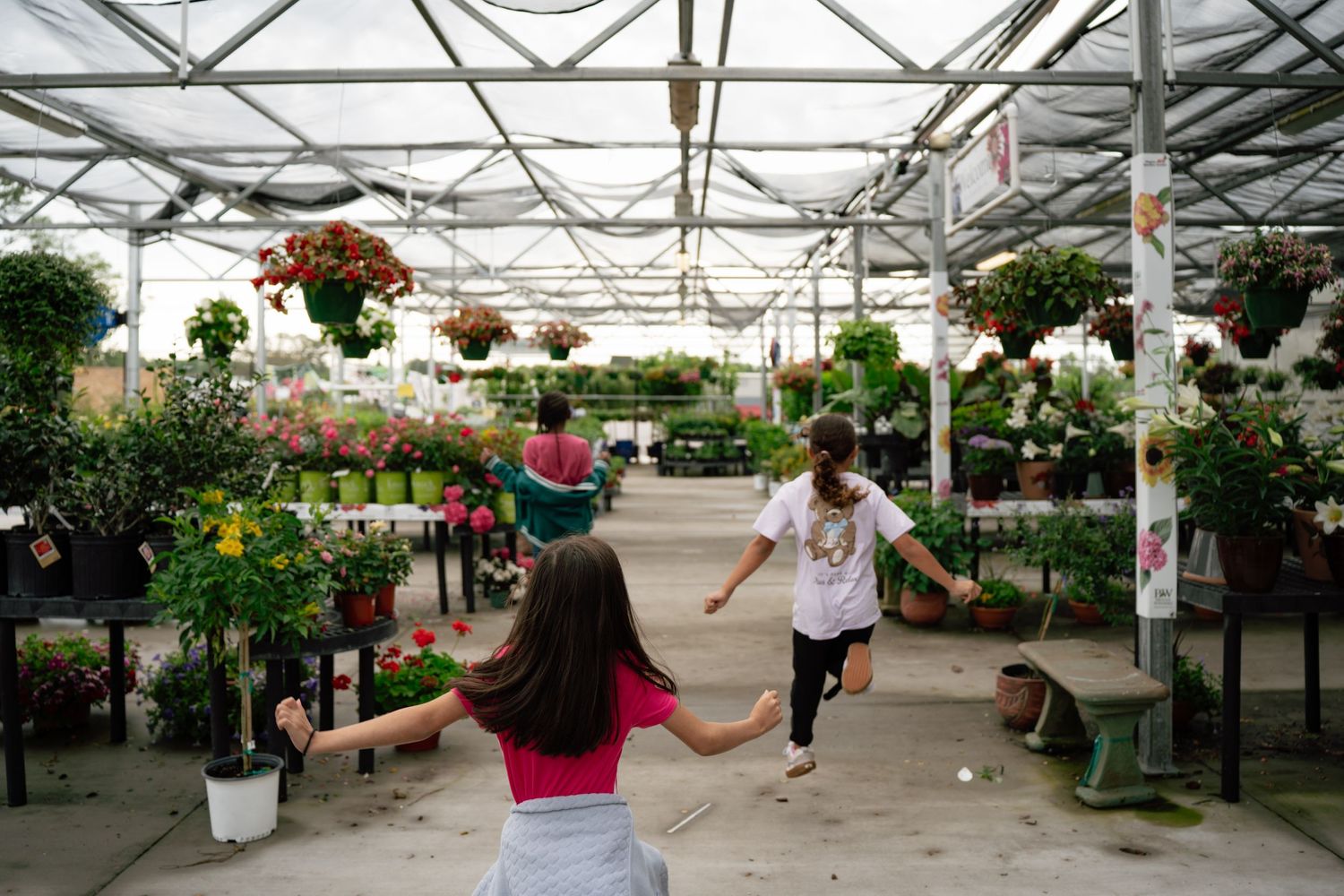 Children playing and running through a colorful greenhouse garden center filled with hanging flowers and plants.