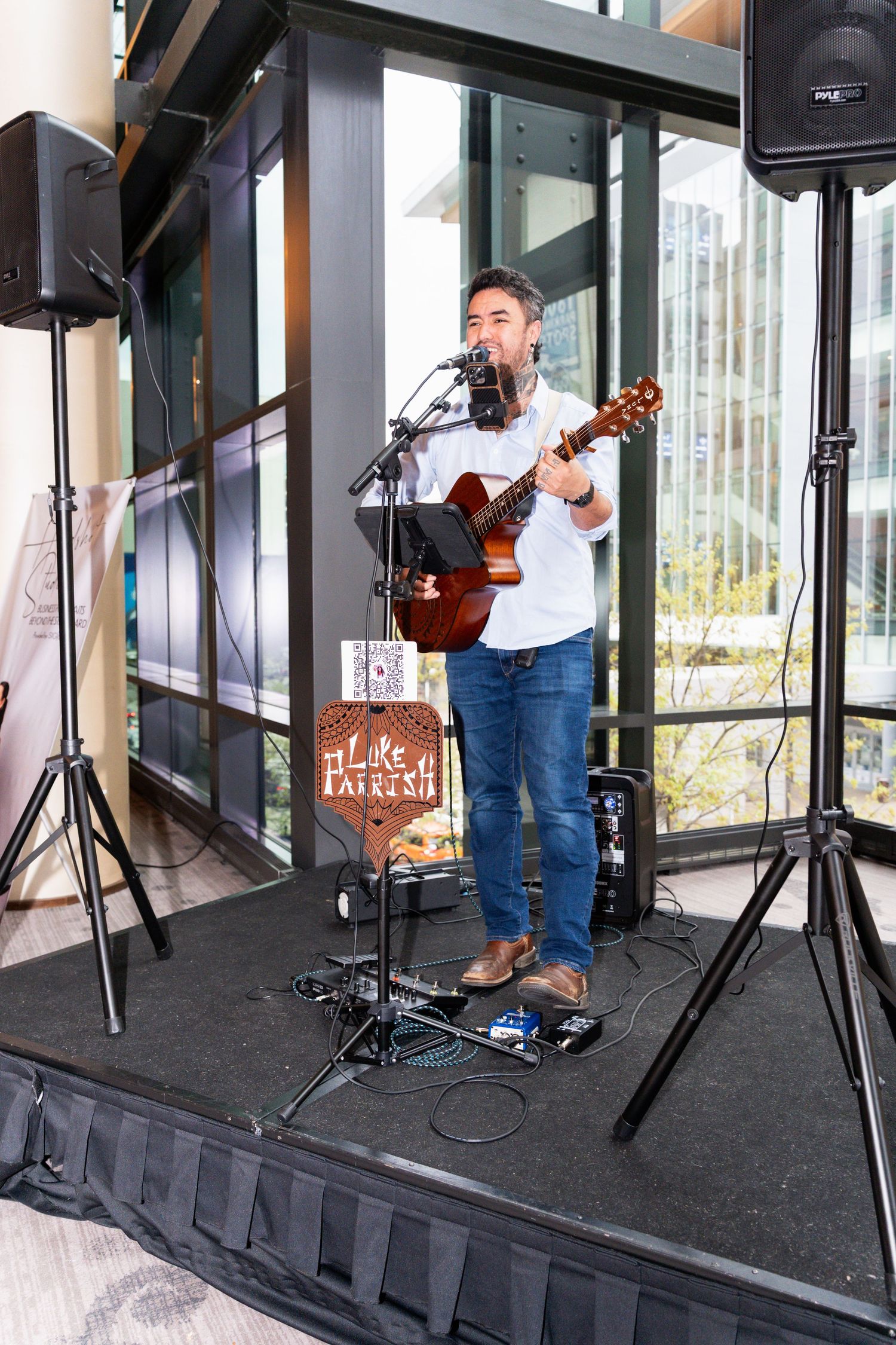 Musician playing acoustic guitar and singing into microphone on small stage with speaker stands at indoor venue.