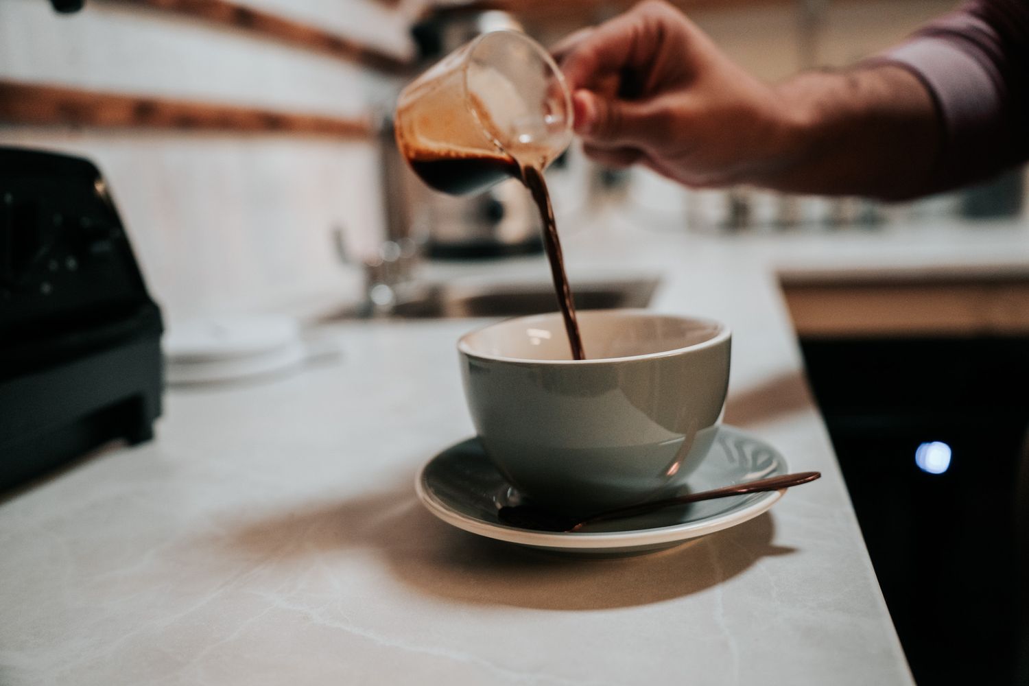 Someone pours a dark liquid from a glass container into a gray ceramic coffee cup on a white countertop.