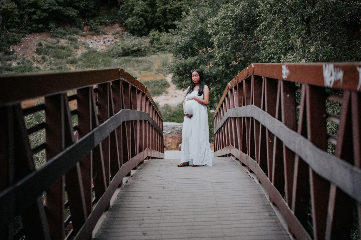 A person in a white dress stands on a rustic wooden bridge surrounded by natural scenery.