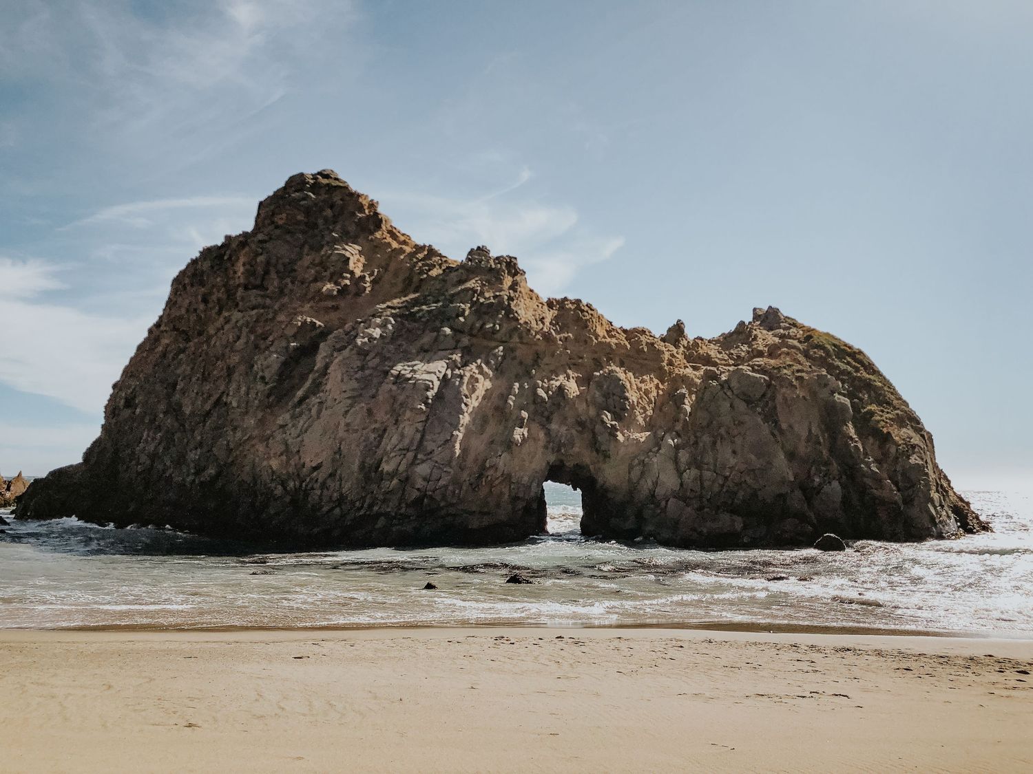 Large rock formation with natural arch opening on sandy beach under cloudy sky at Pfeiffer Beach, Big Sur, California.
