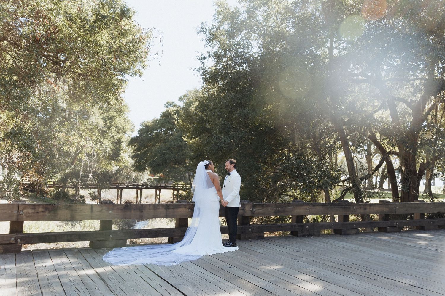 A couple in wedding attire stands together on a wooden deck surrounded by sunlit trees in a park setting.