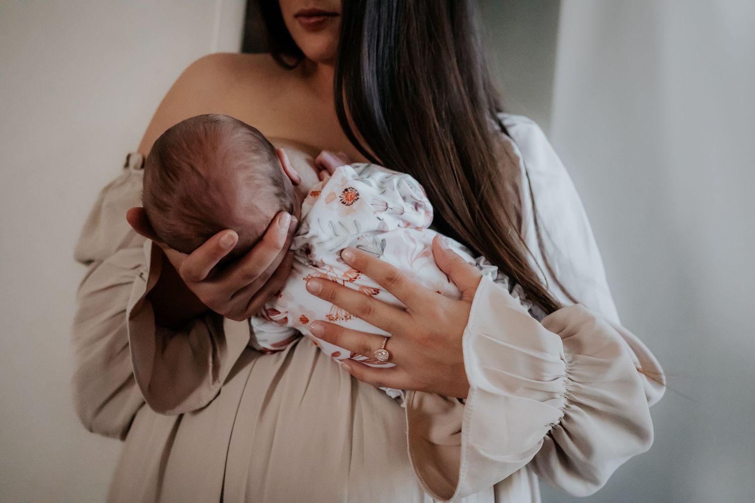 A parent in a beige dress cradles a newborn baby wearing white against a light background.