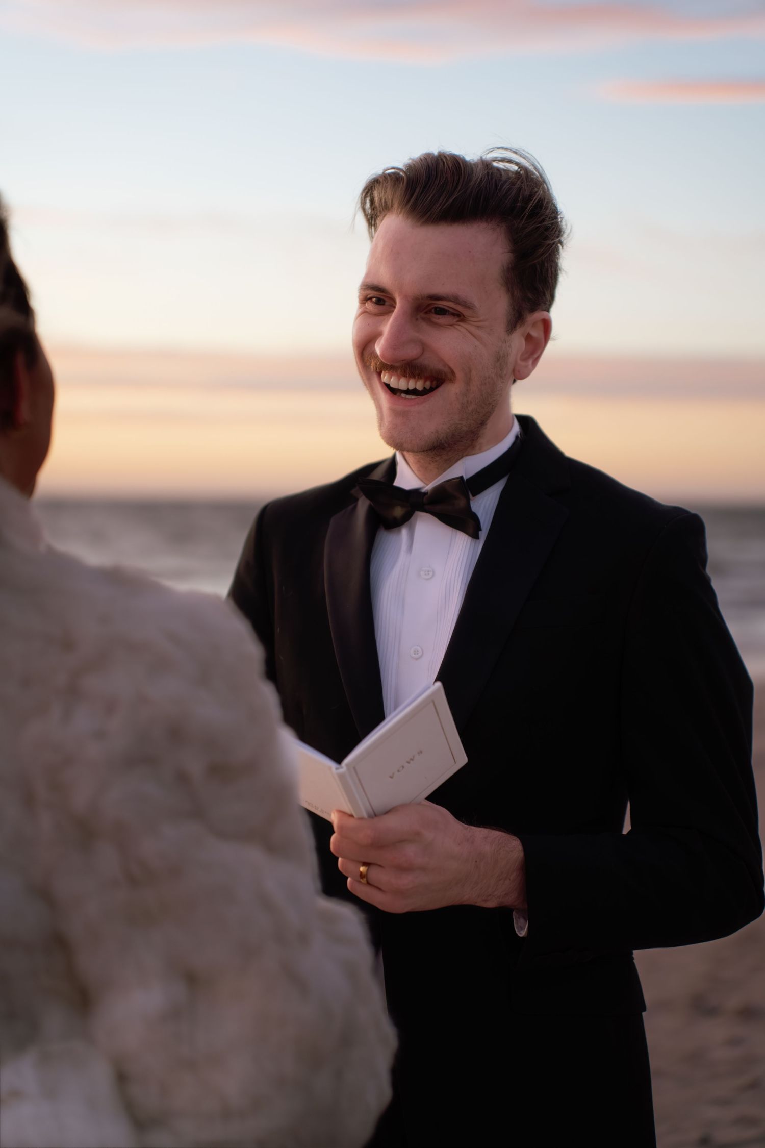 A person in a black tuxedo and bowtie holds a book while smiling against a sunset sky backdrop.