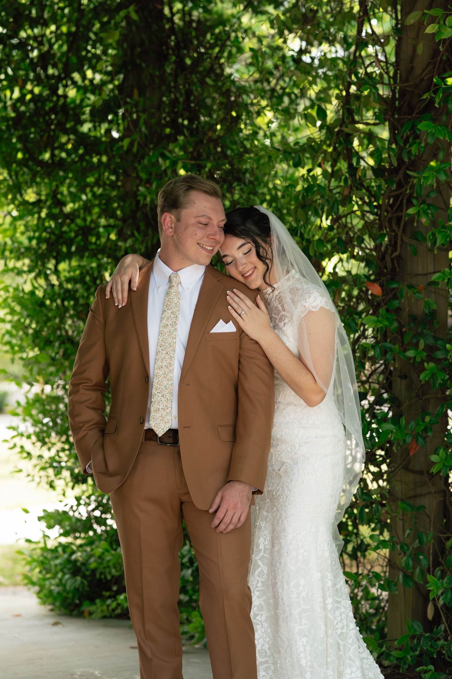 Bride in lace gown and veil leans on groom in brown suit with gold tie, posing under green foliage.