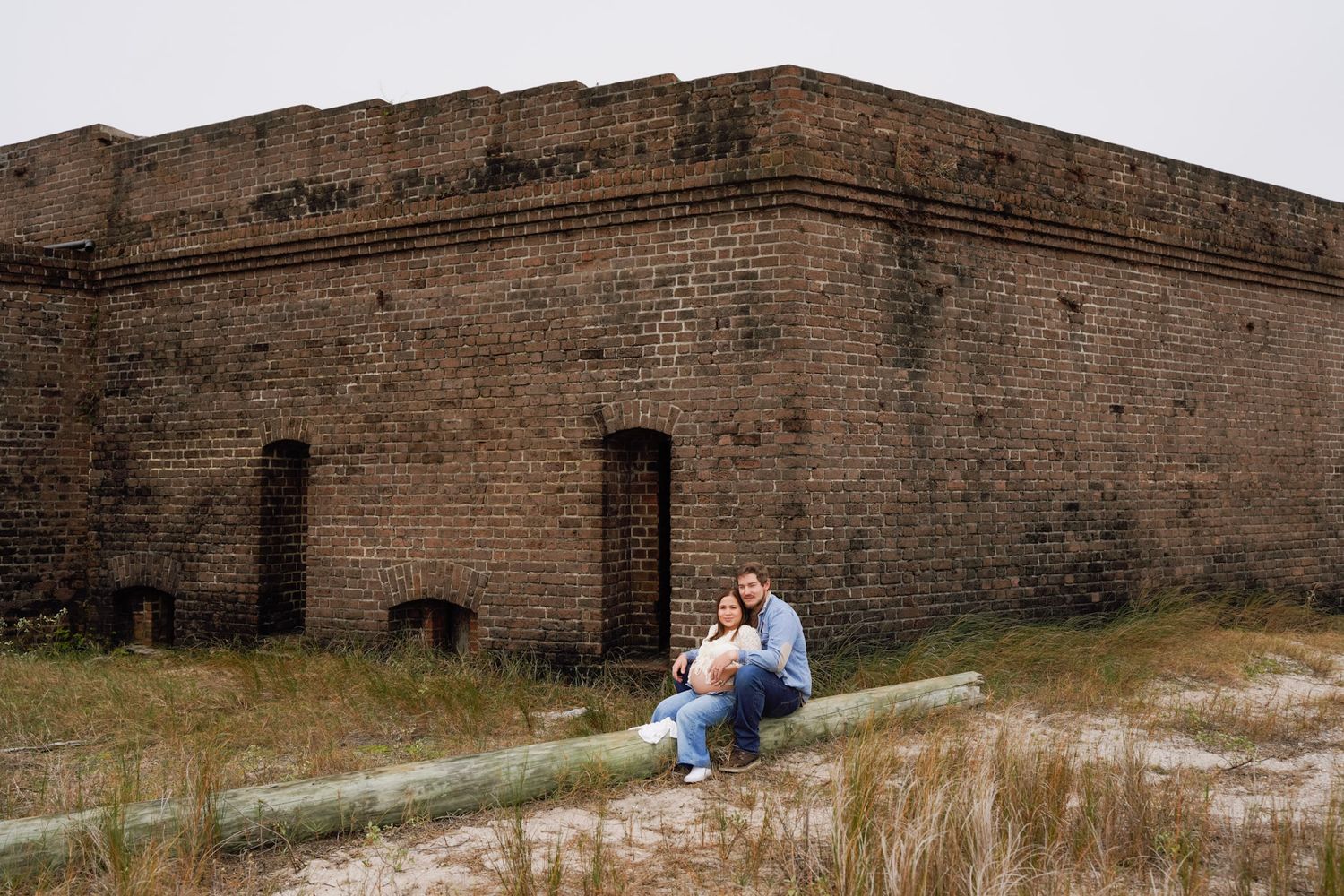 Historic brick fortress walls stretch across a coastal landscape with people relaxing on the sandy ground nearby.