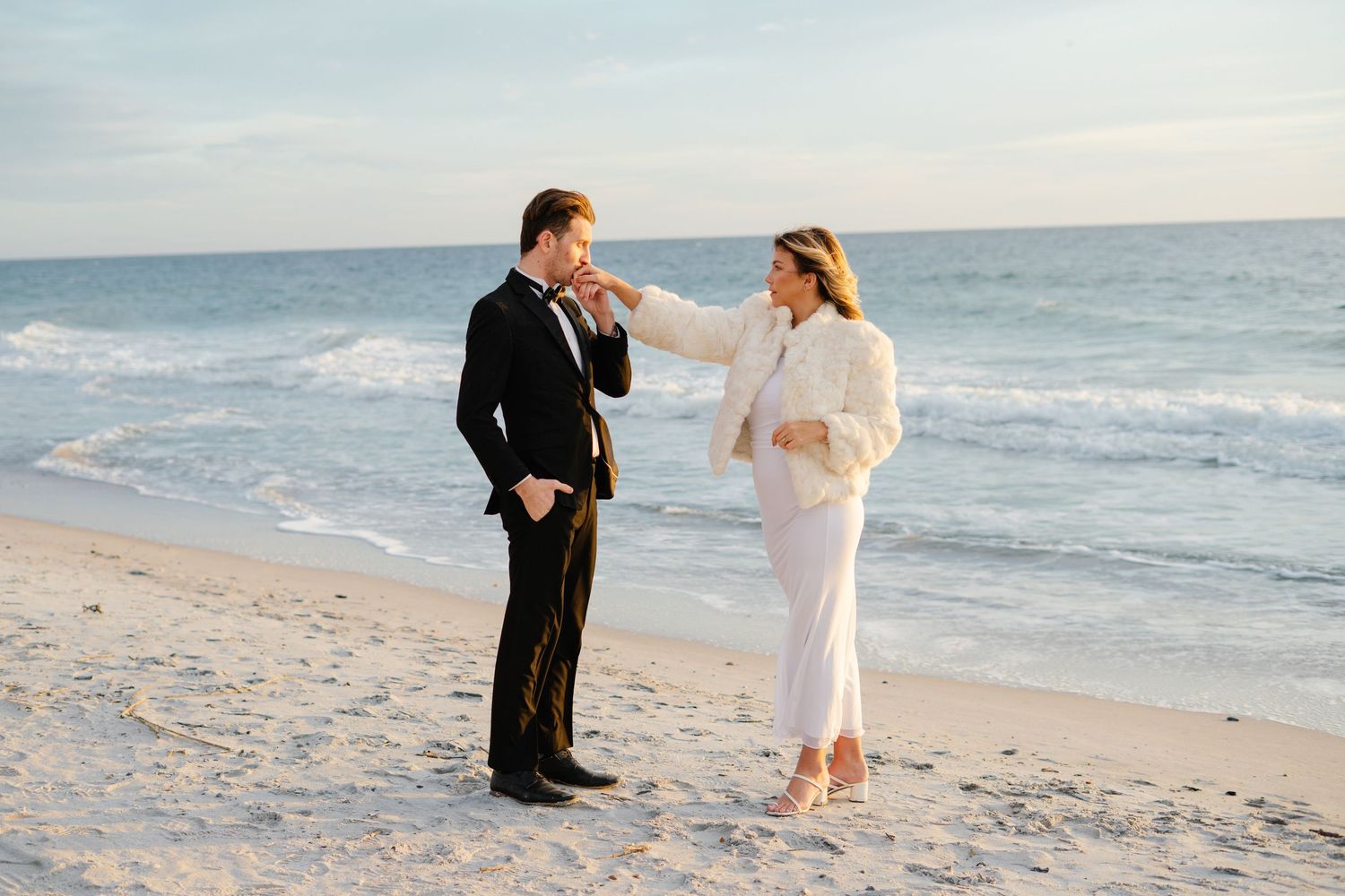 A couple in formal attire shares a romantic moment while walking along a sandy beach at sunset.
