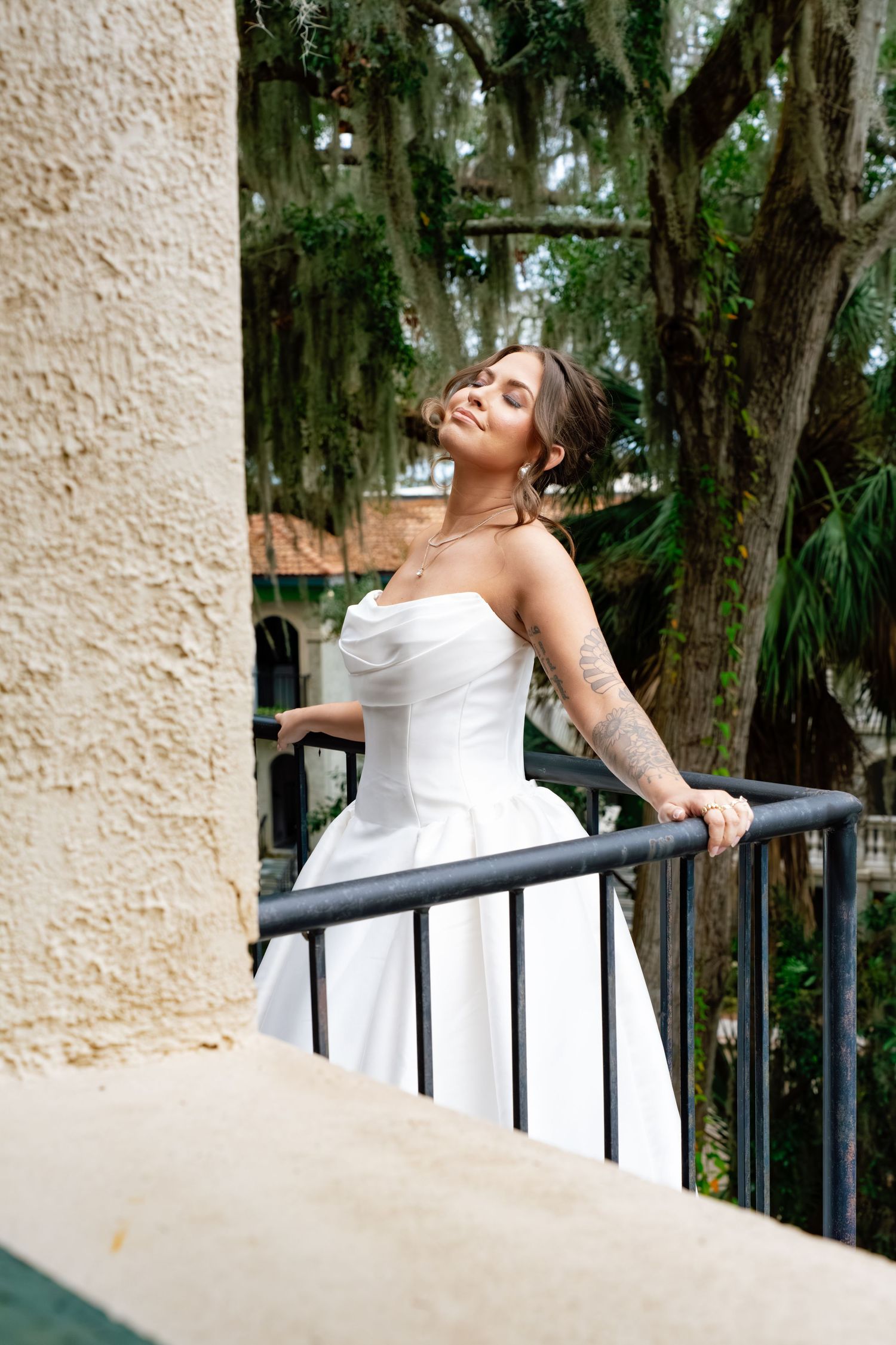 A bride in a white wedding dress leans back against a railing in an outdoor garden setting with Spanish moss trees.