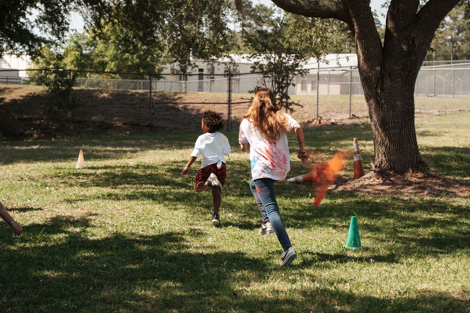 Two children running outdoors on grass near orange smoke cones and a tree during a fun outdoor activity.