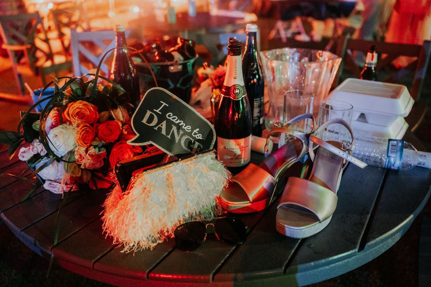 Round table with wedding reception items including a Come Dance sign, wine bottle, floral bouquet, and silver heels.