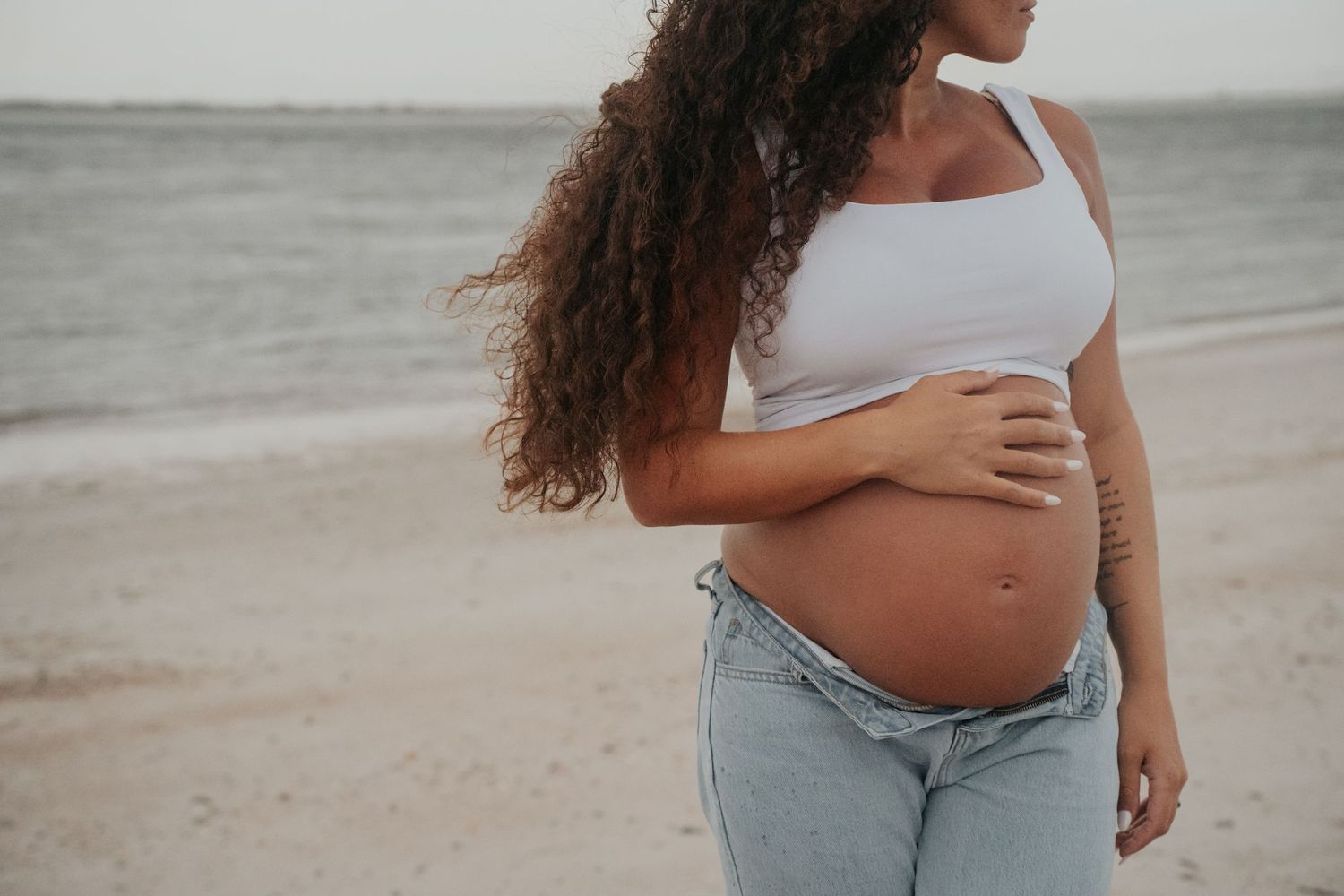 Pregnant woman in white tank top and jeans stands on beach at sunset.