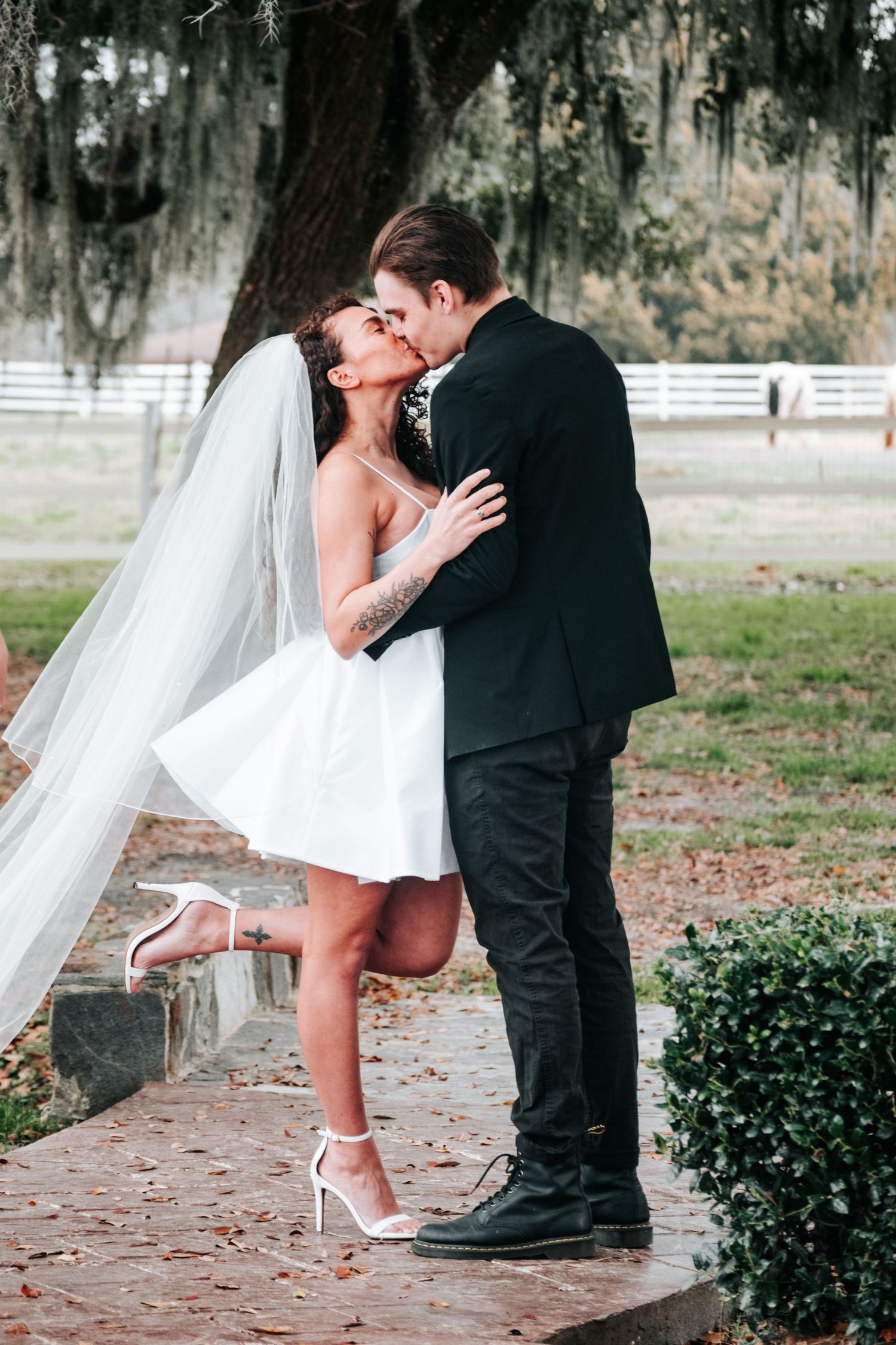A romantic wedding couple shares a kiss on a pathway under Spanish moss trees, with the bride wearing a short white dress and veil.