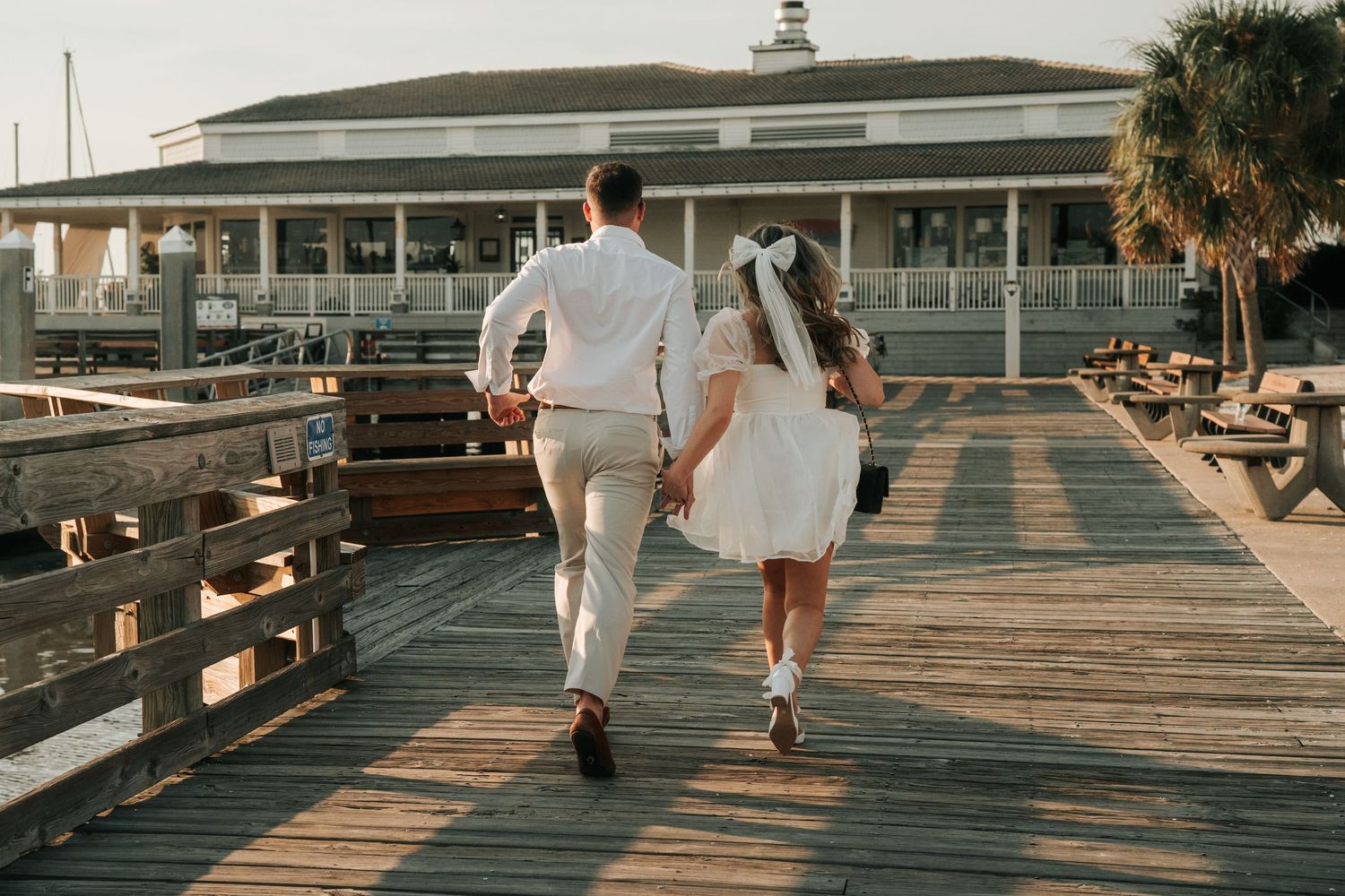 A couple in white outfits walking together on a wooden pier at sunset.
