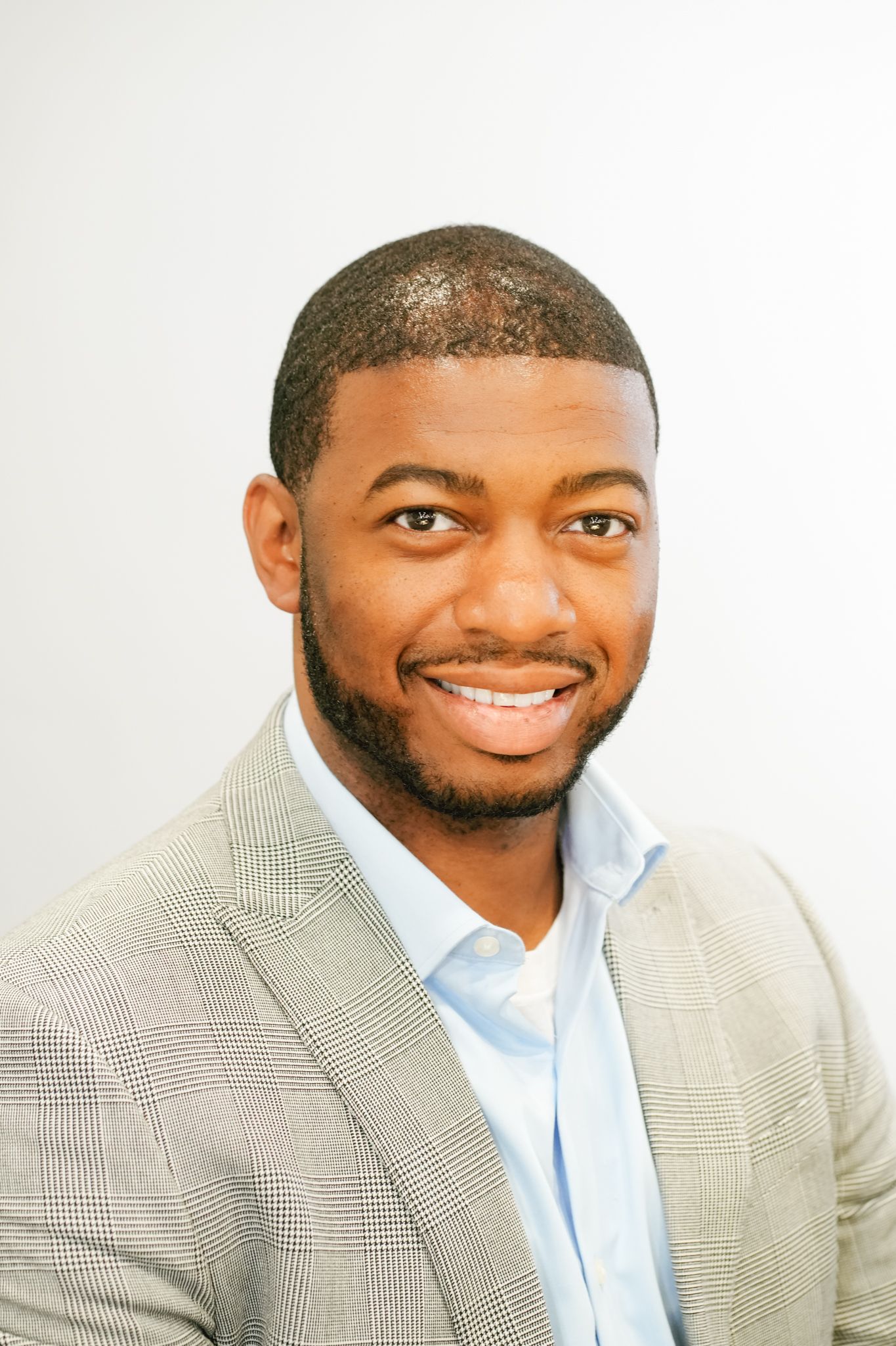 Smiling young Black man in a gray plaid blazer and light blue shirt poses for a professional headshot photo.