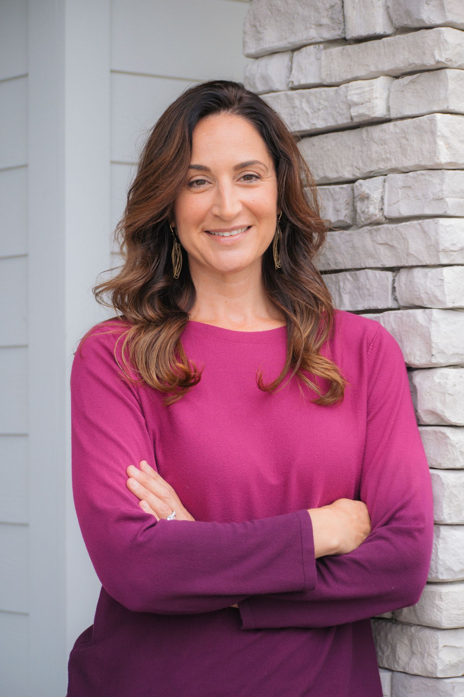 A smiling woman with brown hair in a magenta top stands with arms crossed against a stone wall backdrop.