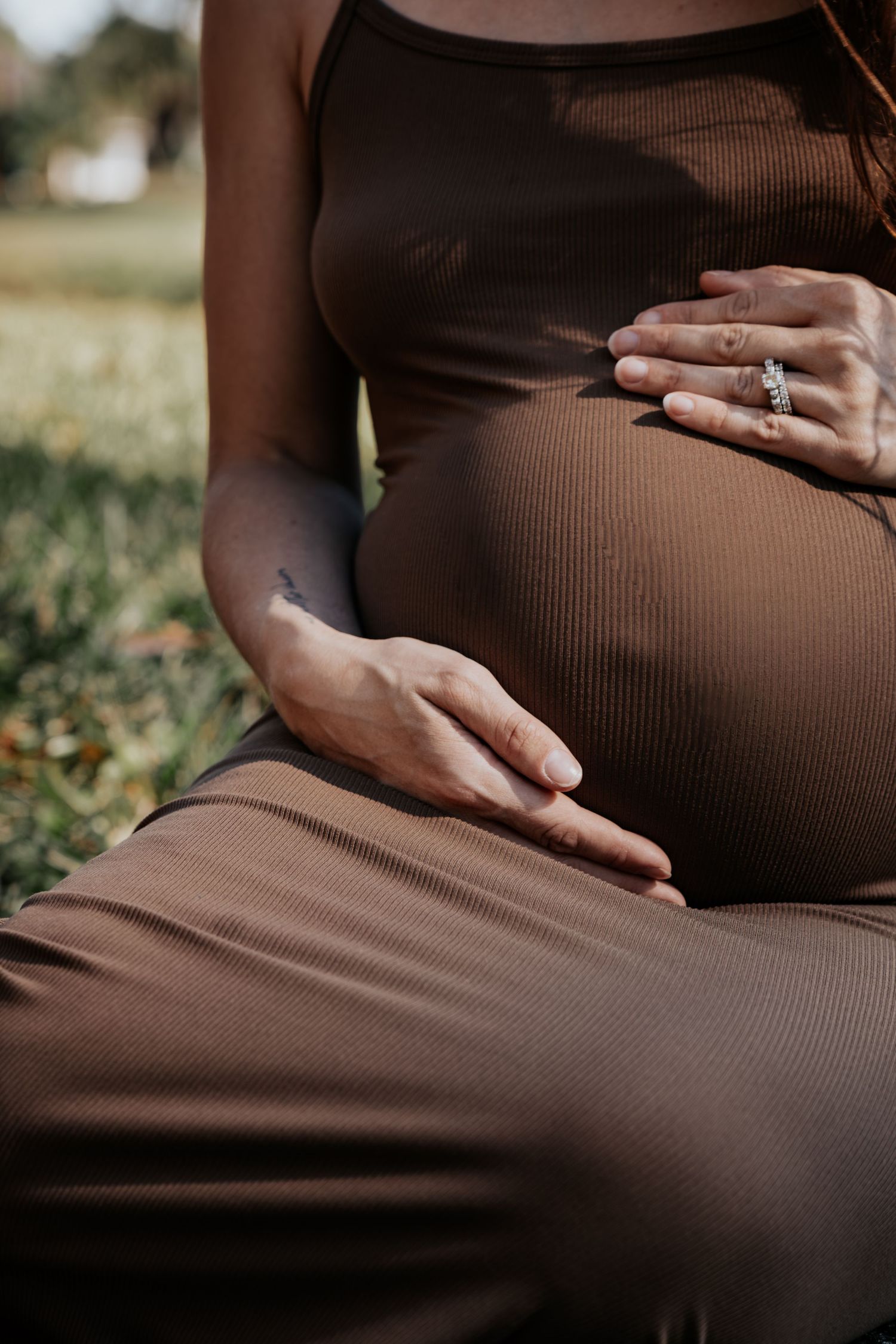 Pregnant woman in brown ribbed dress cradling baby bump outdoors, wearing diamond ring, sitting in sunlit garden.