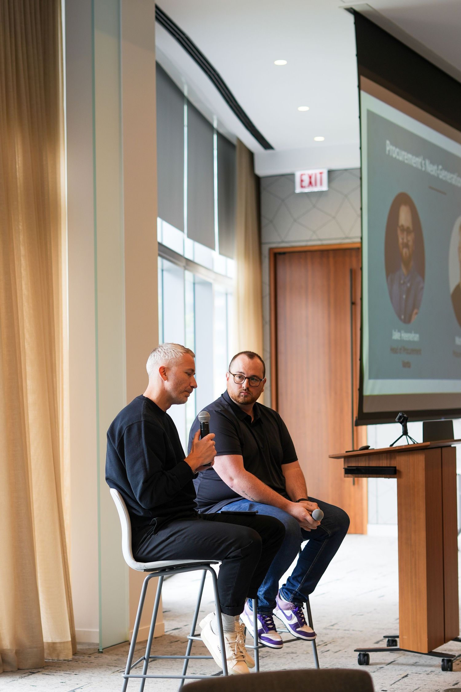 Two men seated on chairs engage in conversation during a professional presentation event with a projection screen behind them.
