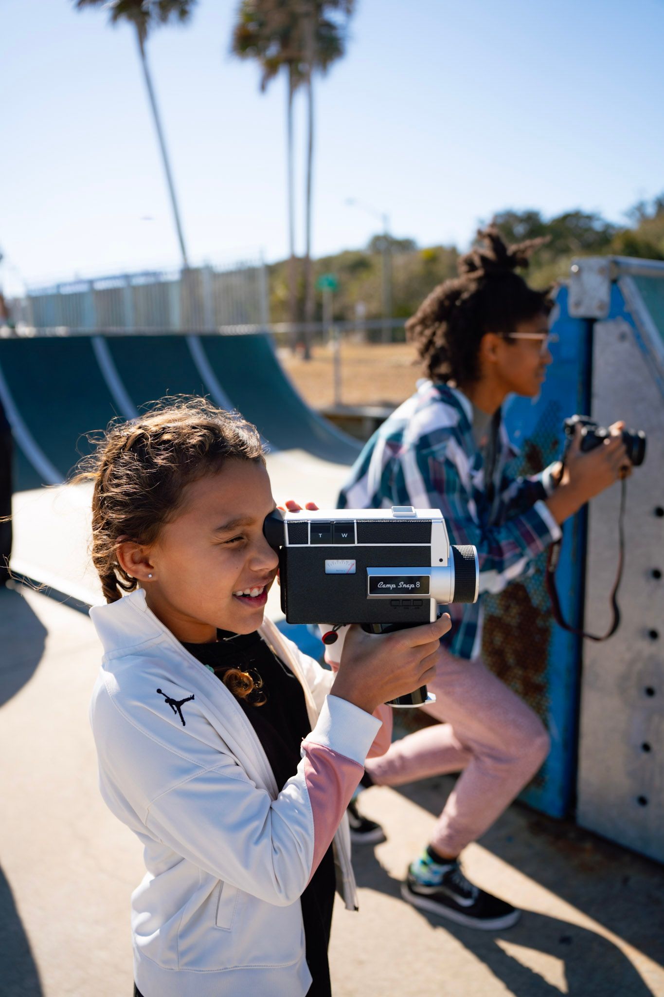 A young girl films with a vintage Super 8 camera at a skate park on a sunny day, with a woman holding a camera behind her.
