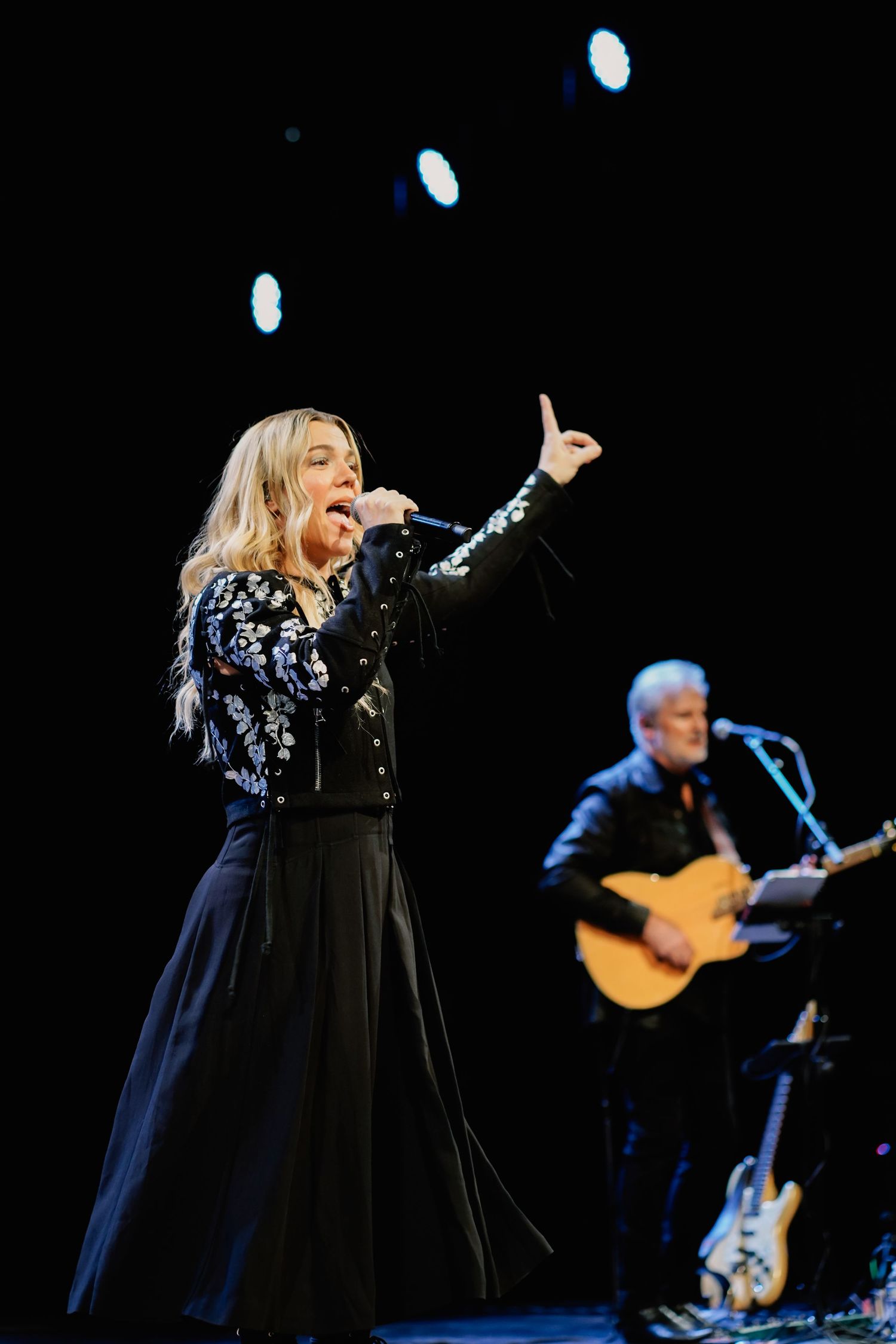 Female singer performing on stage in black floral dress, pointing upward with guitarist in background under stage lights.