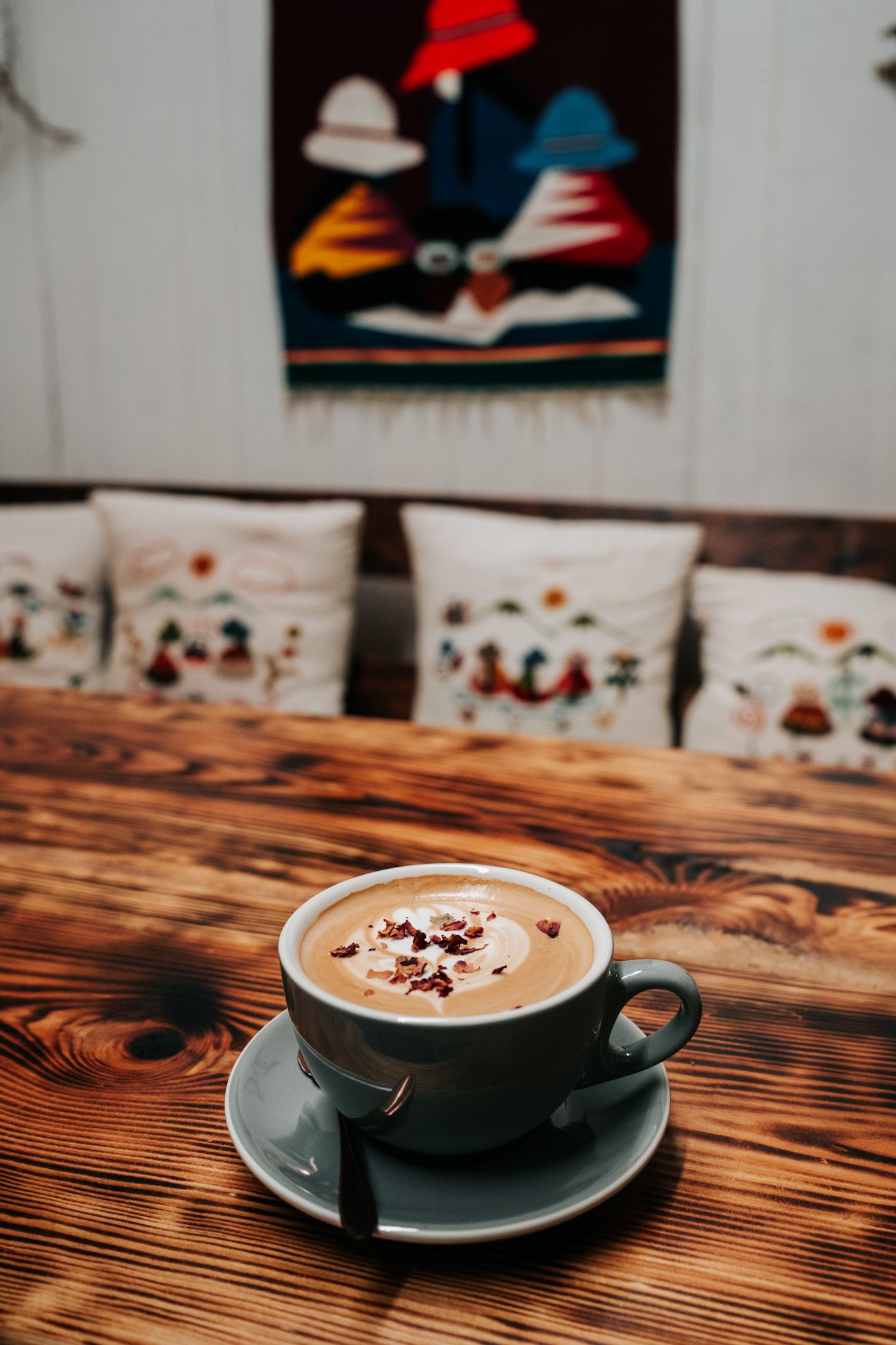 A cup of hot chocolate with chocolate sprinkles on a wooden table, with decorative pillows and wall art in background.