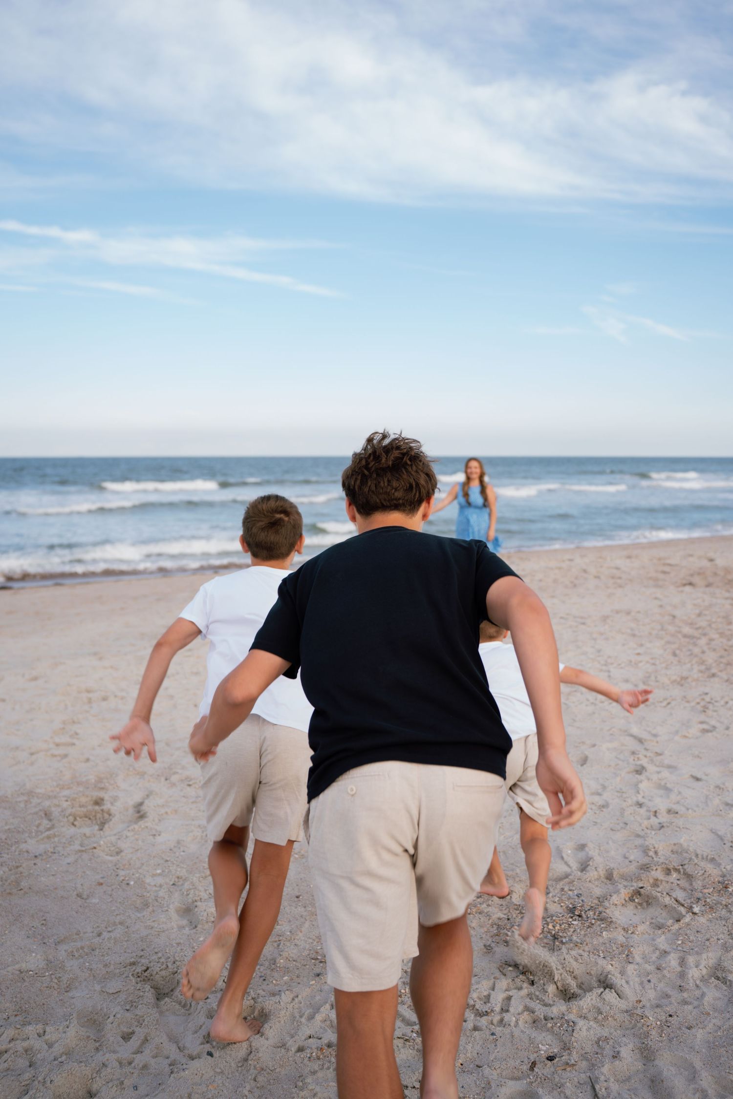 Children running toward a woman on a sandy beach with ocean waves and blue sky in the background.