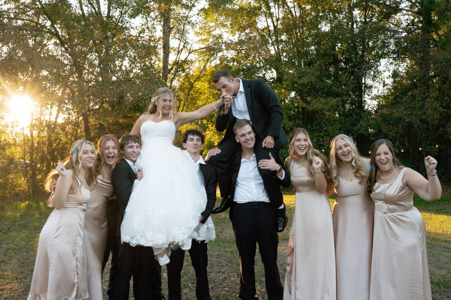 Wedding party in champagne colored dresses and black tuxedos pose together during golden hour sunset outdoors.