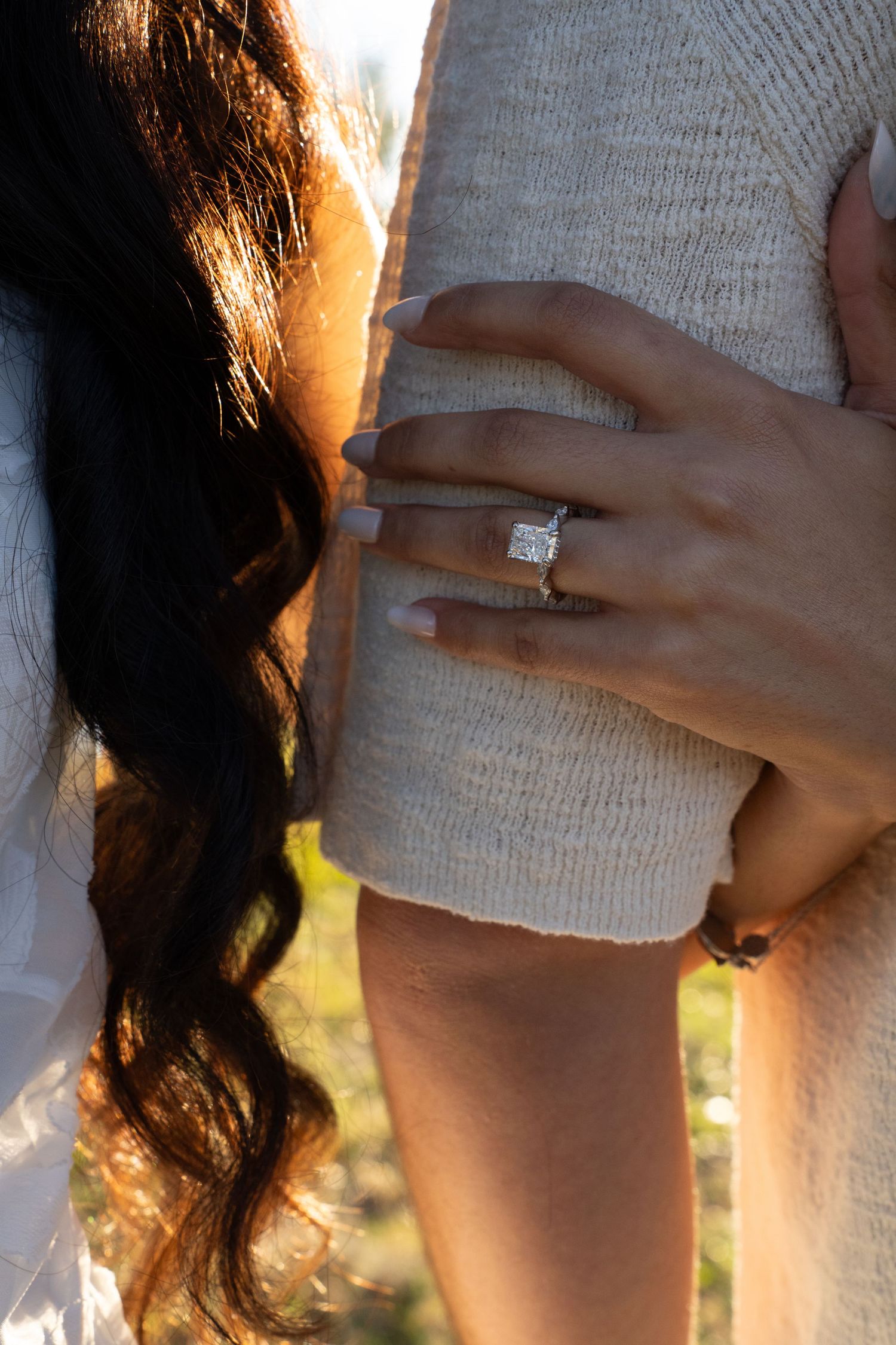 Close-up view of a sparkling diamond engagement ring on a woman's hand against a white knit sweater in sunlight.