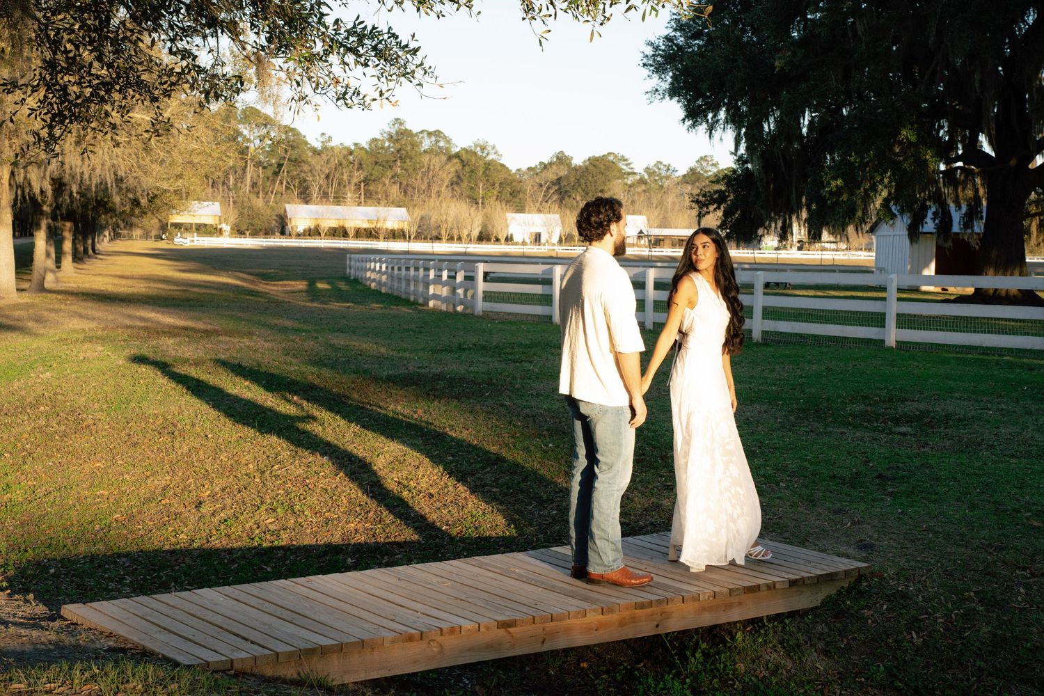 A couple walks hand in hand on a wooden boardwalk surrounded by trees and white fencing during golden hour.