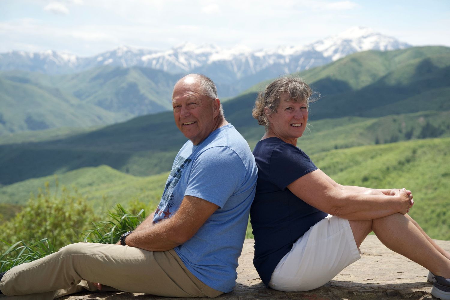 Two people sit back-to-back outdoors enjoying a sunny mountain vista with snow-capped peaks in the background in Utah