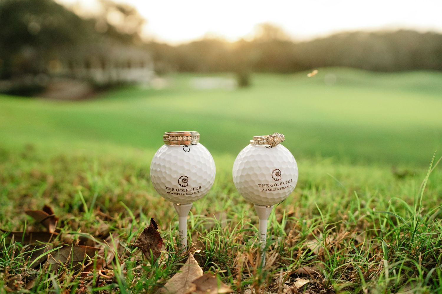 Two wedding rings are displayed on top of golf balls on a grassy golf course during sunset.