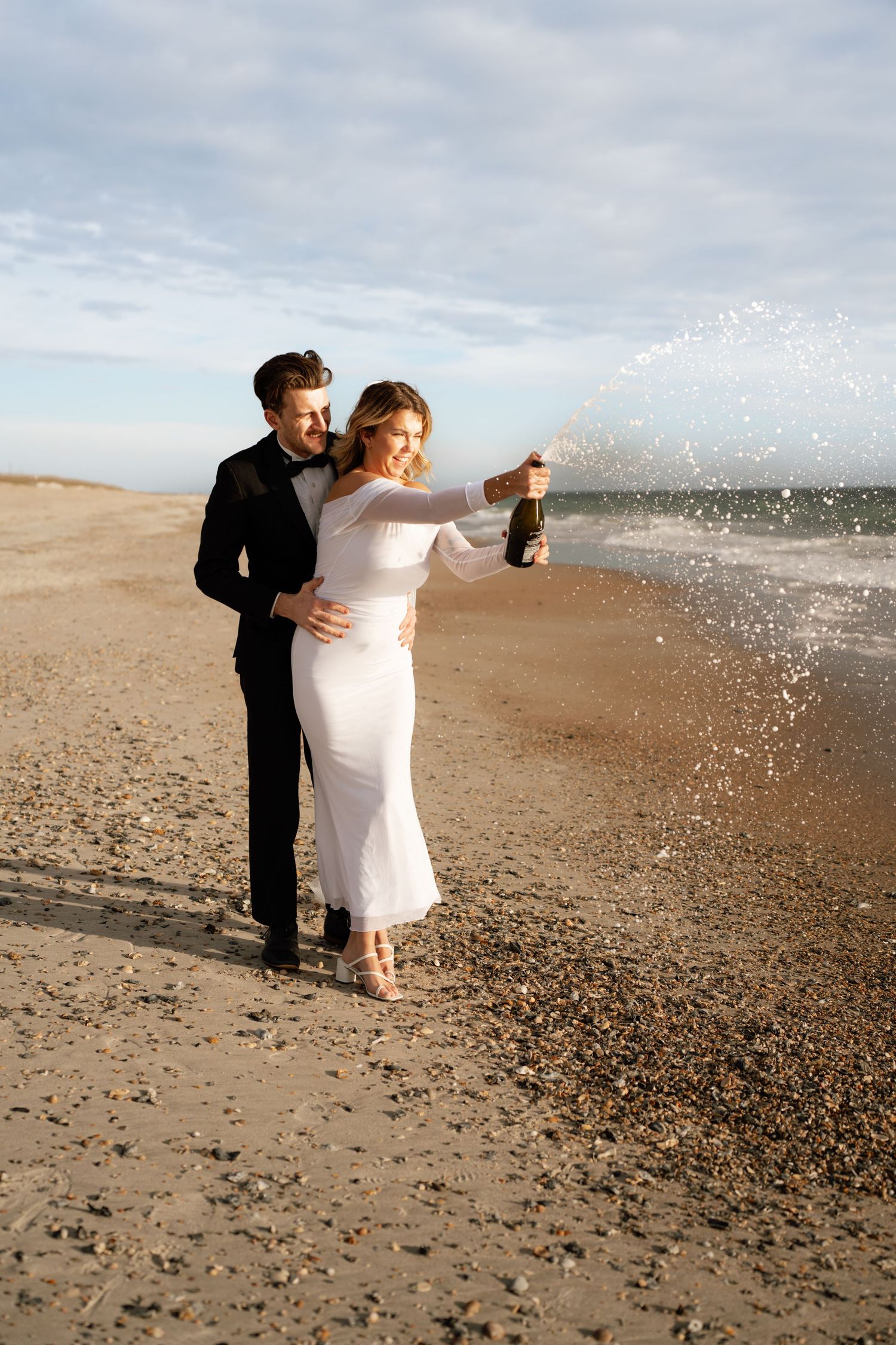 A couple in formal attire celebrating with champagne on a sandy beach at sunset.