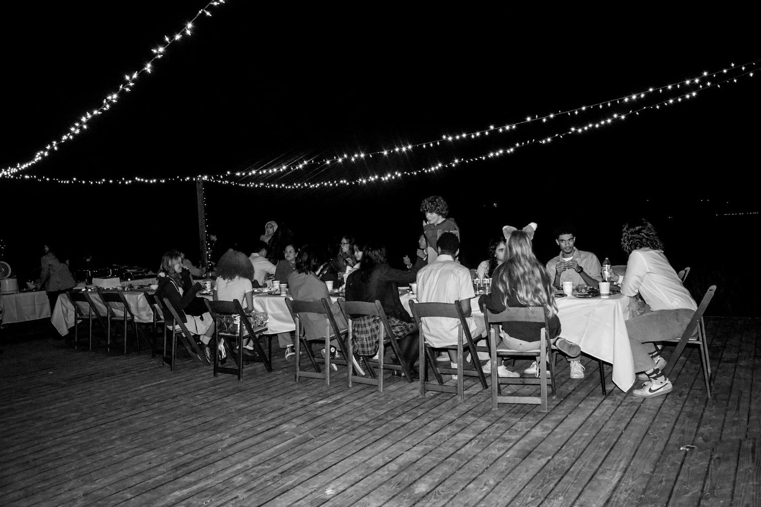 Black and white photo of guests dining outdoors at night under string lights on a wooden deck.