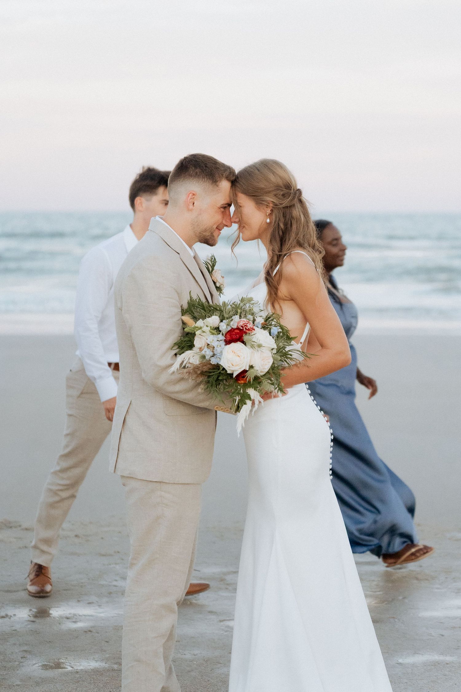 A romantic beach wedding scene with a couple sharing a tender moment in light beige and white attire by the ocean.