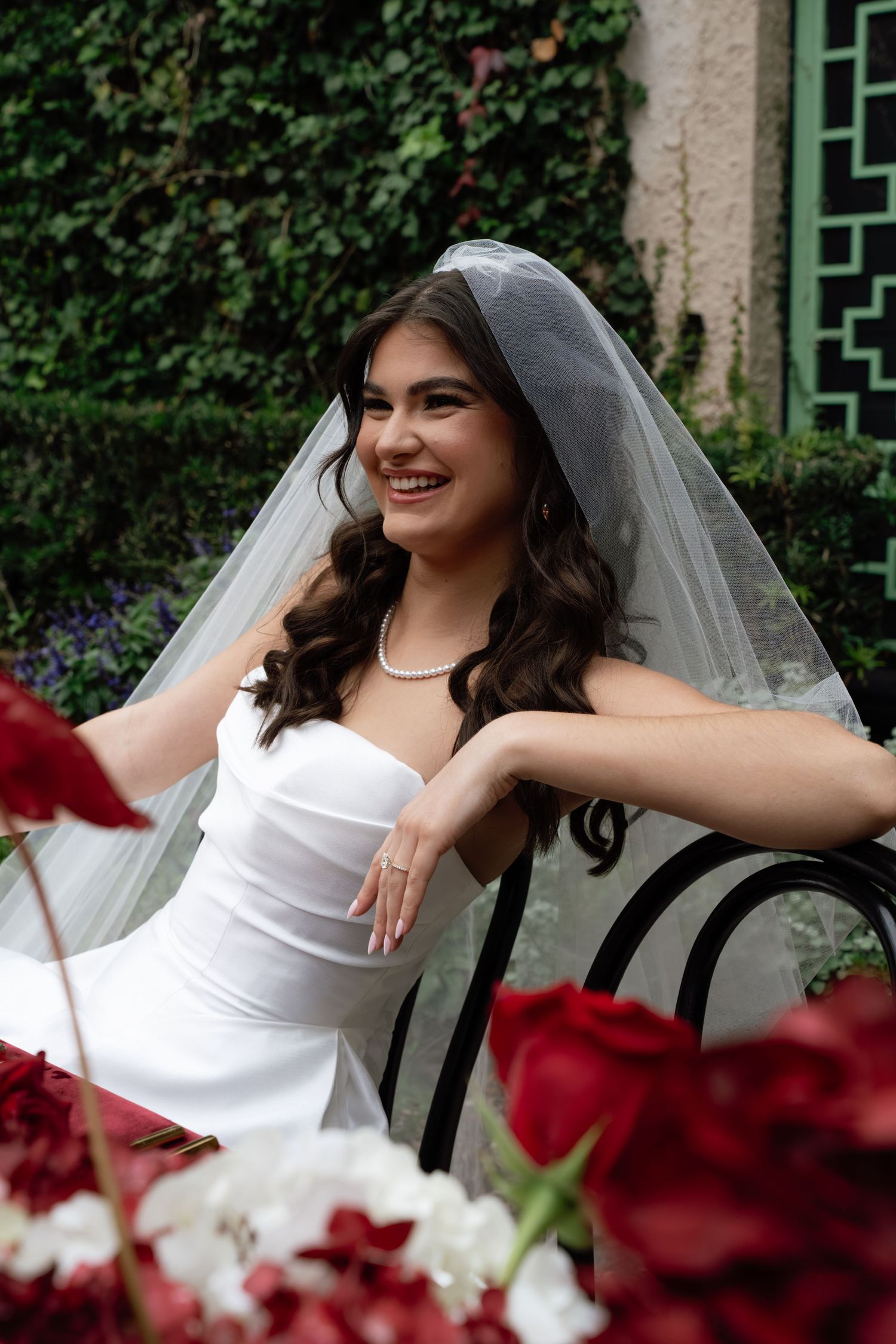 A bride in a white dress poses gracefully on a black bench with red and white flowers in the foreground.