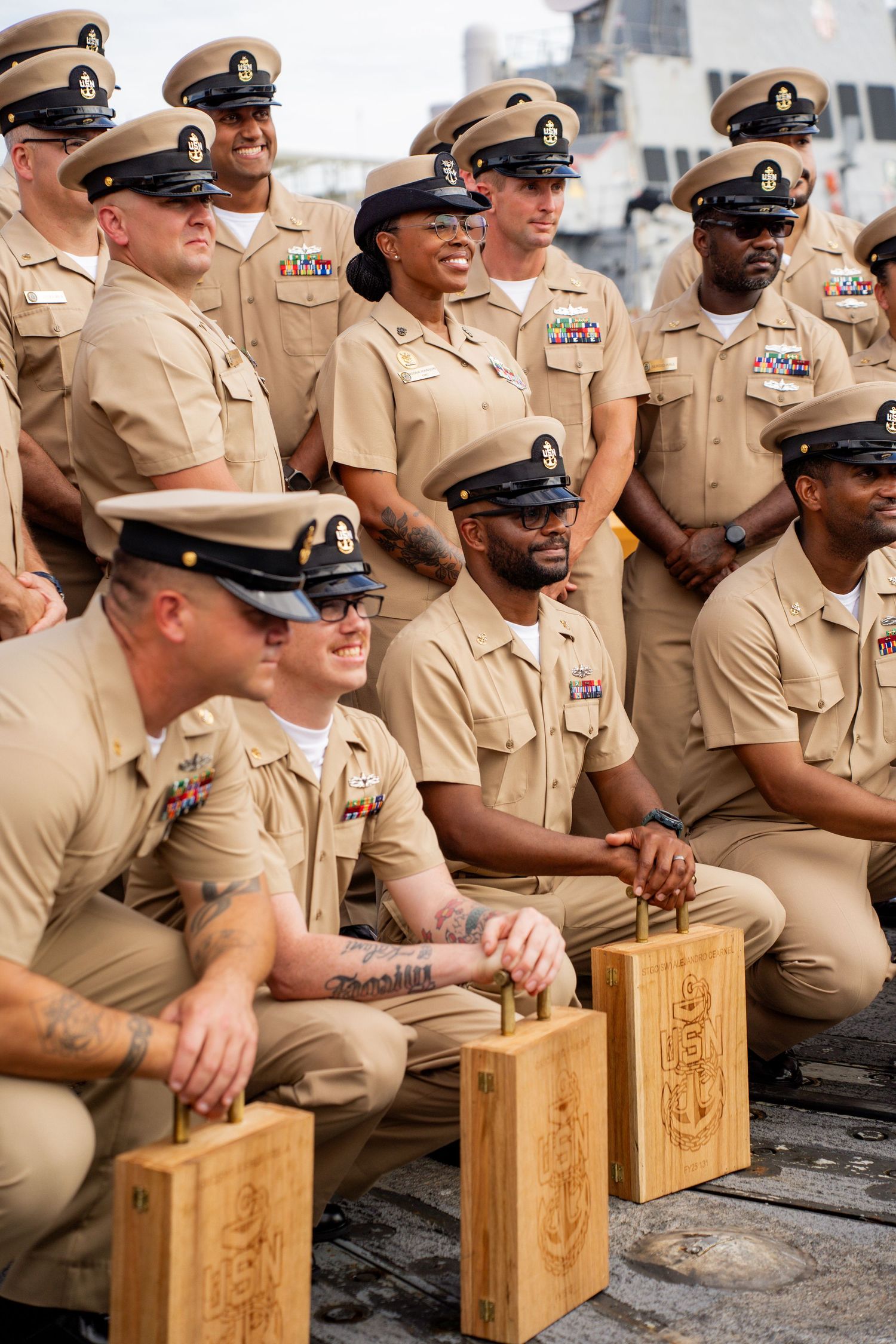A group of U.S. Navy service members in khaki uniforms sit together for a group photo with wooden commemorative plaques.