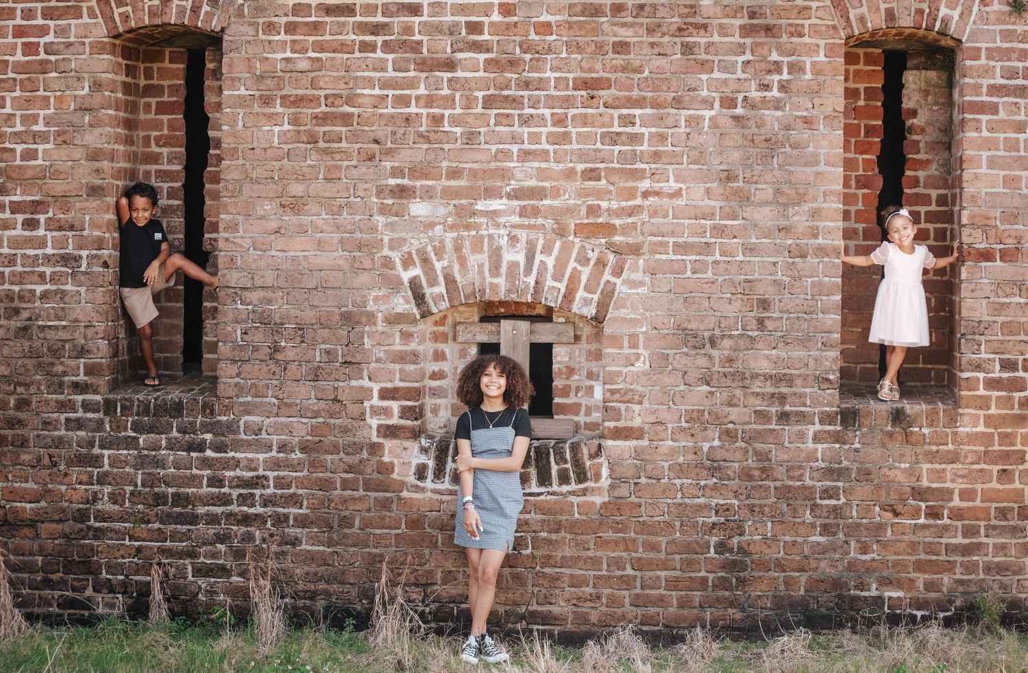 Three kids pose playfully in arched recesses of an old rustic brick wall outdoors at Fort Clinch State Park