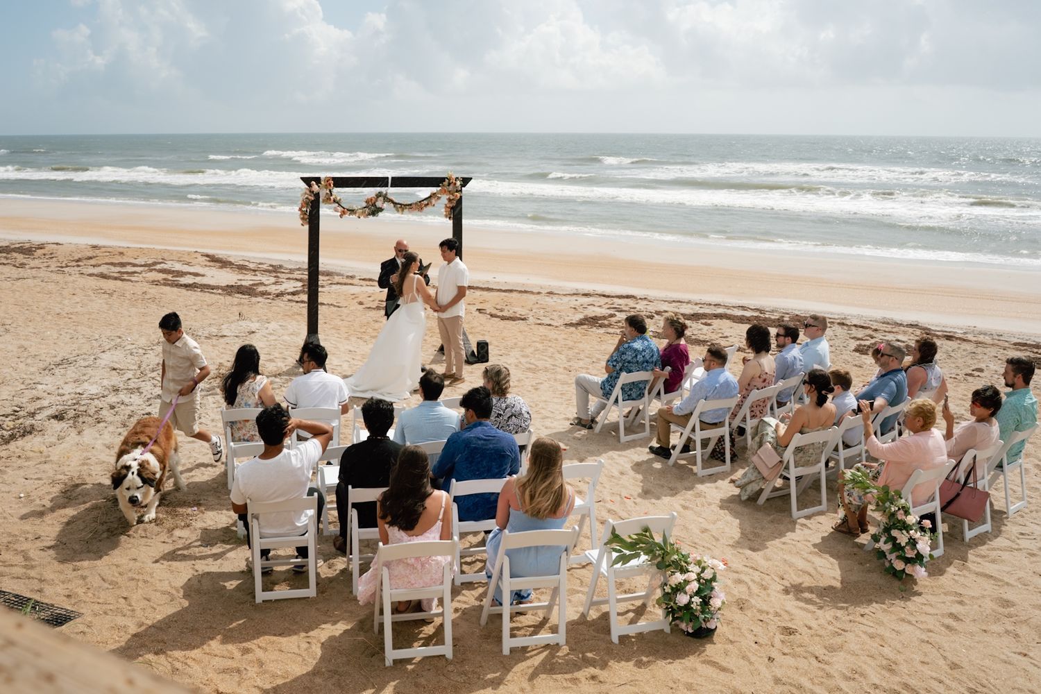 Romantic beach wedding ceremony with couple at wooden arch, guests seated on white chairs by the ocean on sunny sandy shore.