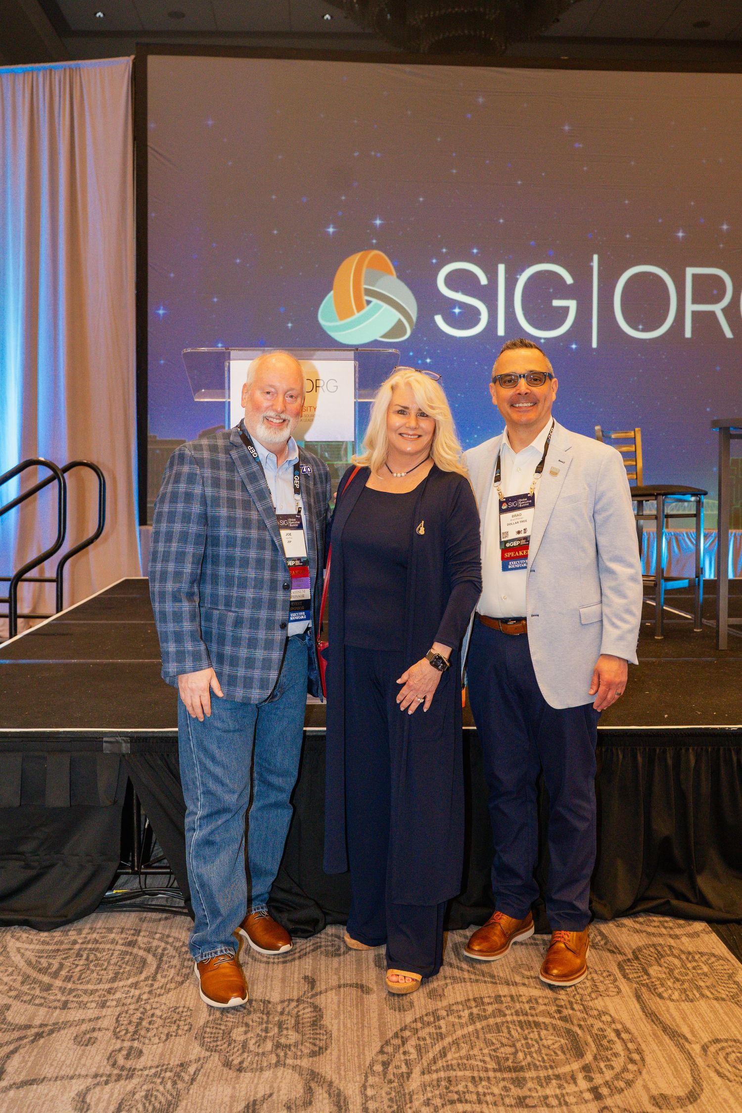 Three professionals pose together smiling in front of a SIG organization event stage backdrop.
