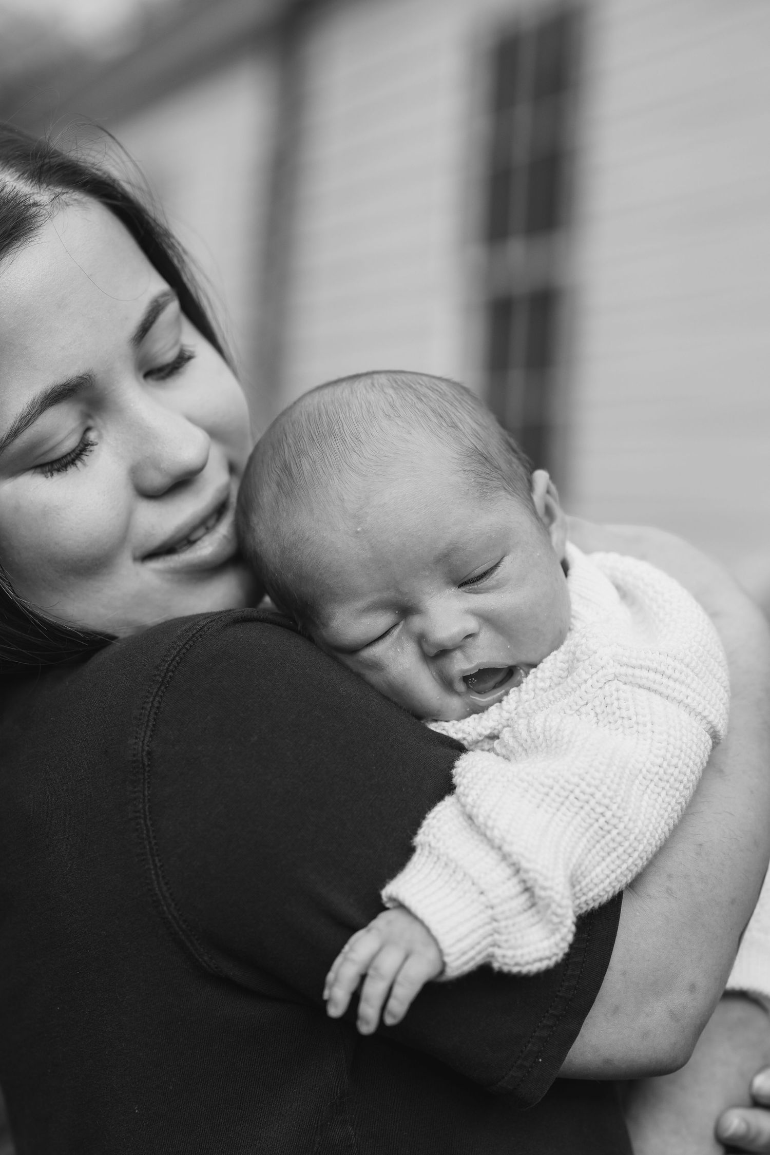 Black and white photo of a mother tenderly holding her sleeping newborn baby close to her chest outdoors.