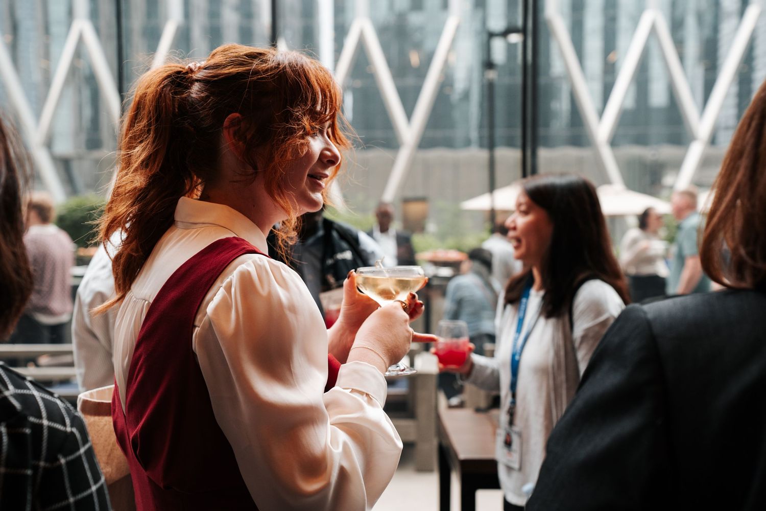 Woman laughing at a corporate networking event, holding a cocktail glass in a modern glass building.