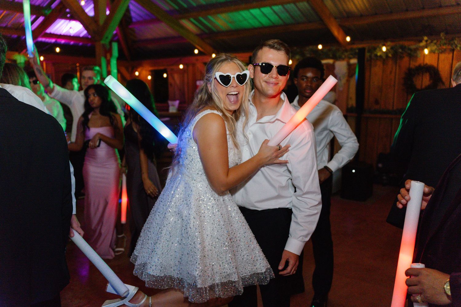 Wedding party guests dance and celebrate on a colorfully lit dance floor in a rustic barn venue.