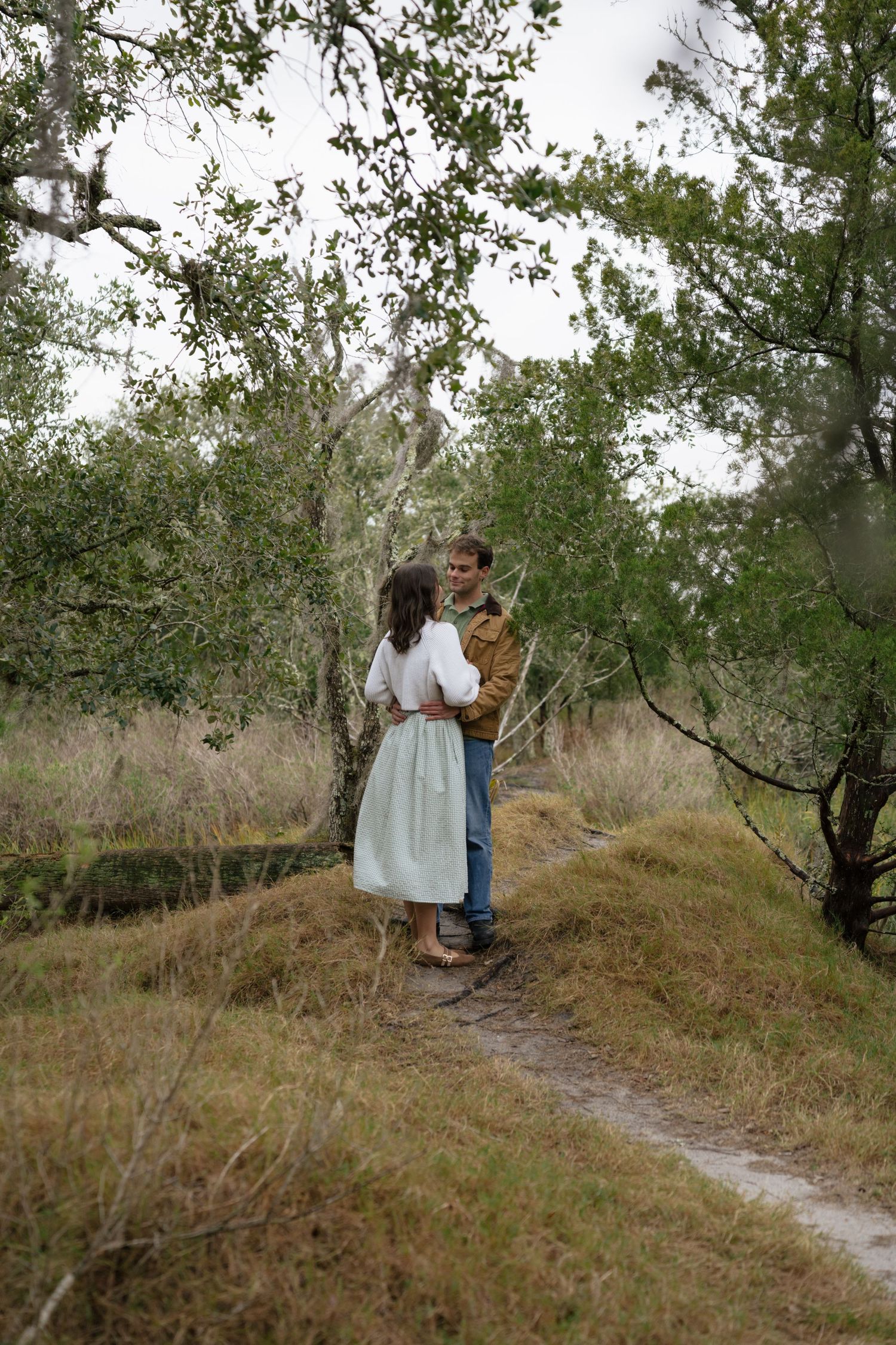 A couple walks together along a winding dirt path through a grove of olive trees on a cloudy day.