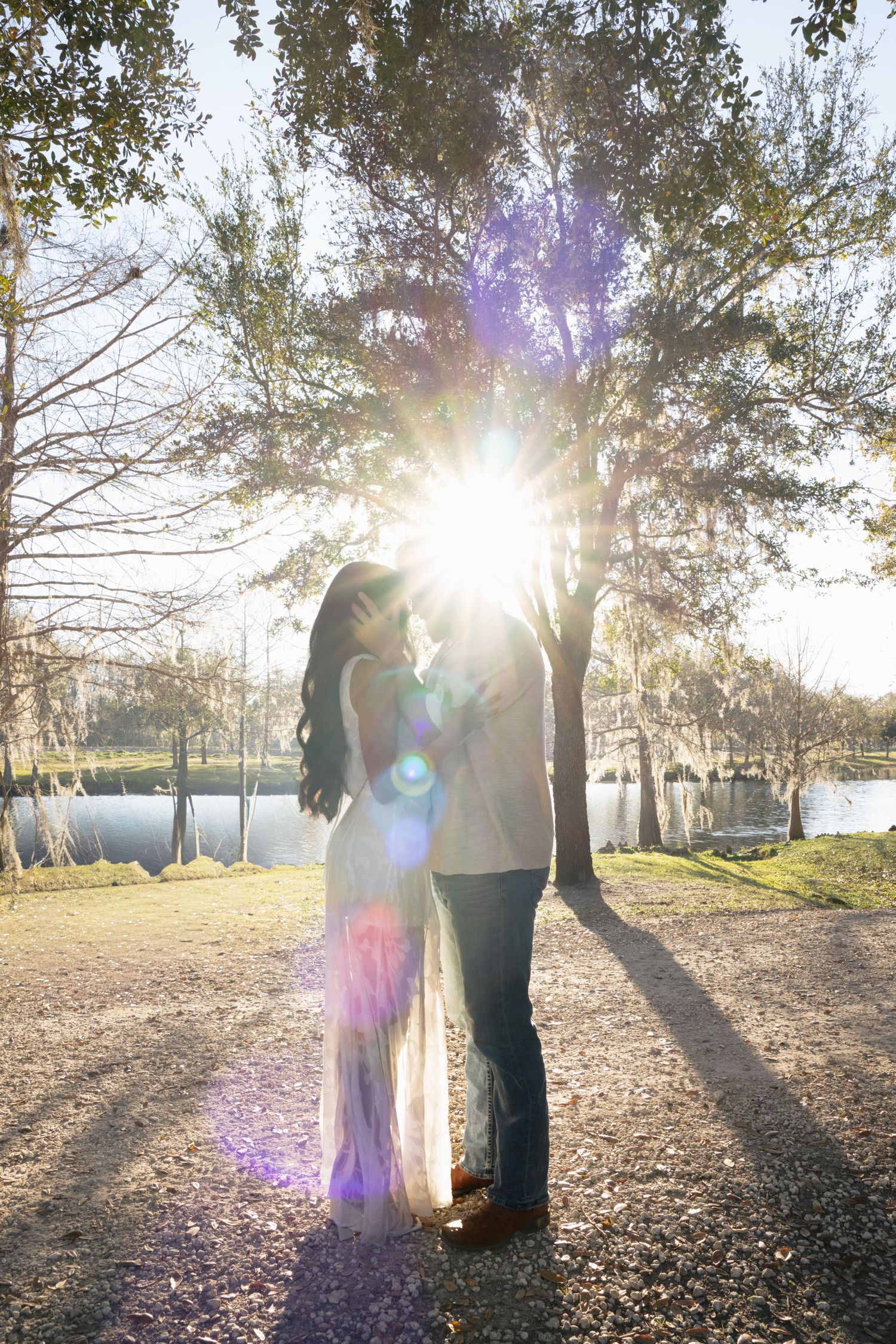 A couple embraces in a romantic sunset moment under a tree by a lake with beautiful lens flare effects.