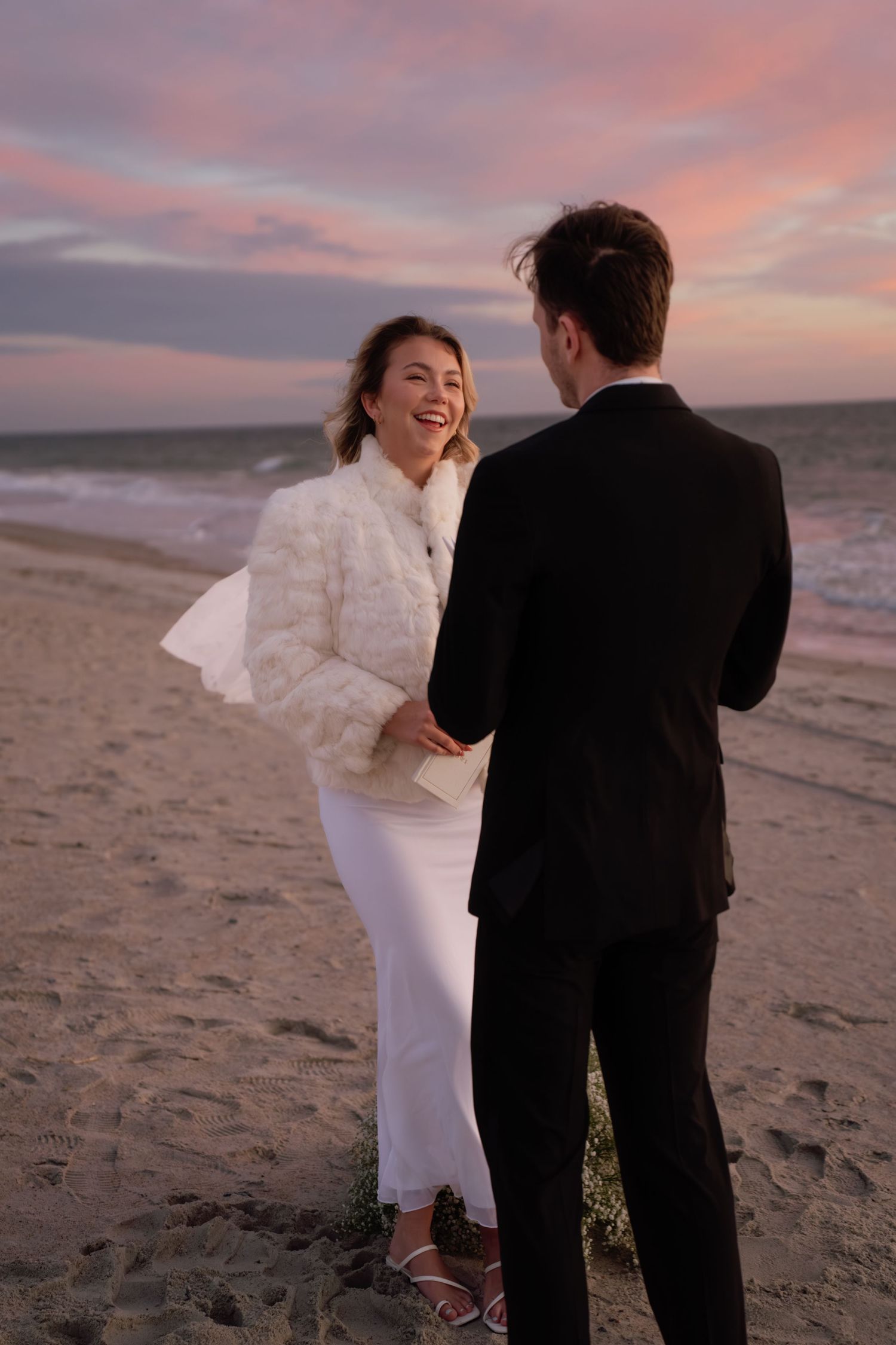 A couple shares a joyful moment on the beach at sunset, with the bride wearing a white dress and fuzzy jacket.
