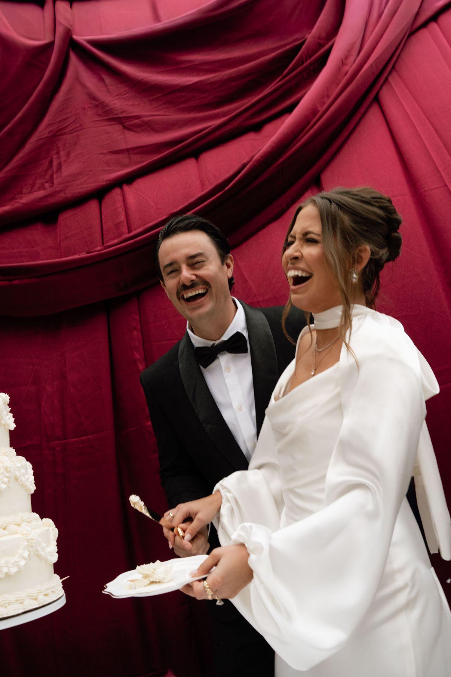 A bride and groom share a joyful moment while cutting their wedding cake against a deep red backdrop.
